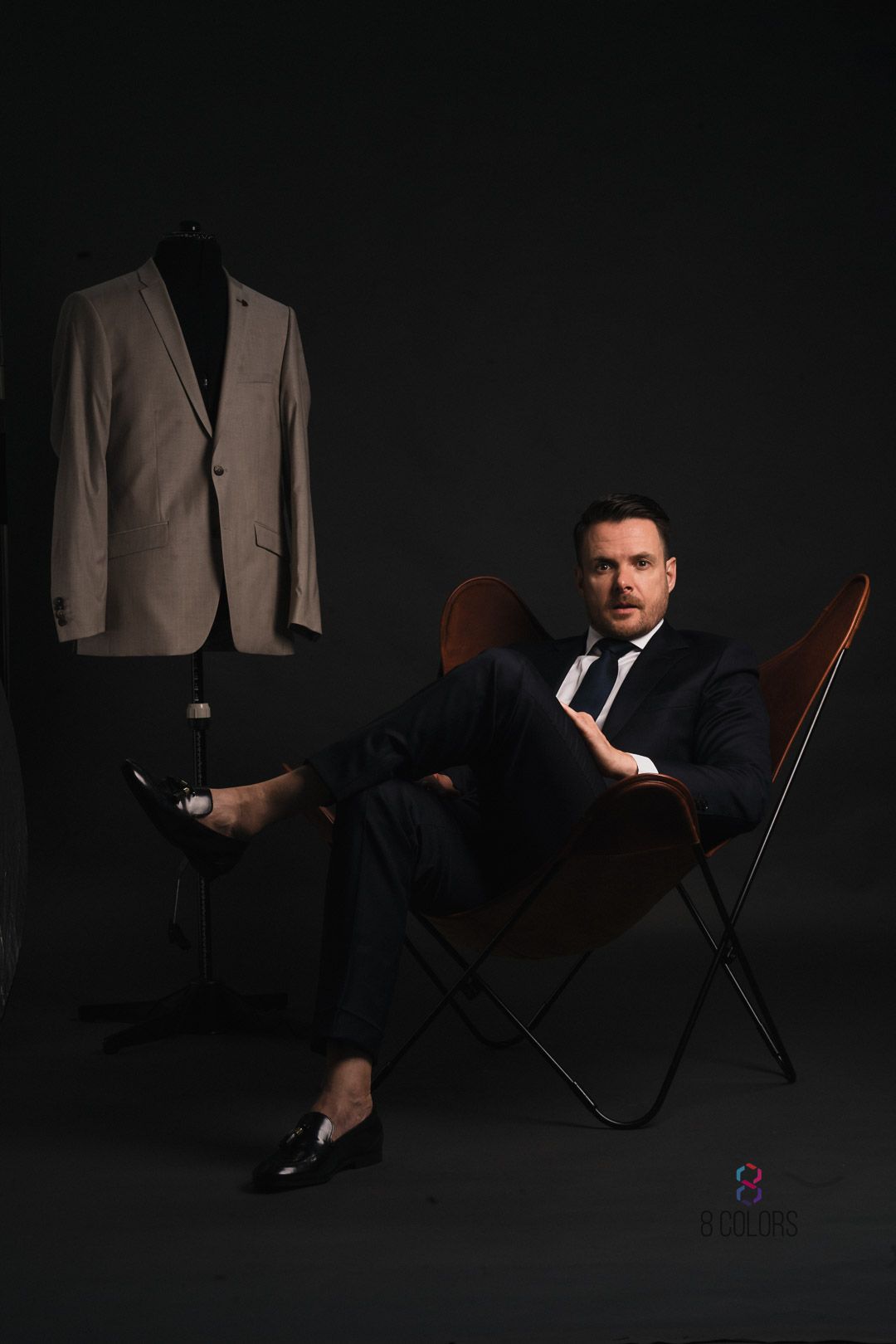 Dark moody fashion photo with man seated in leather chair and tan suit jacket displayed on wall against black background.