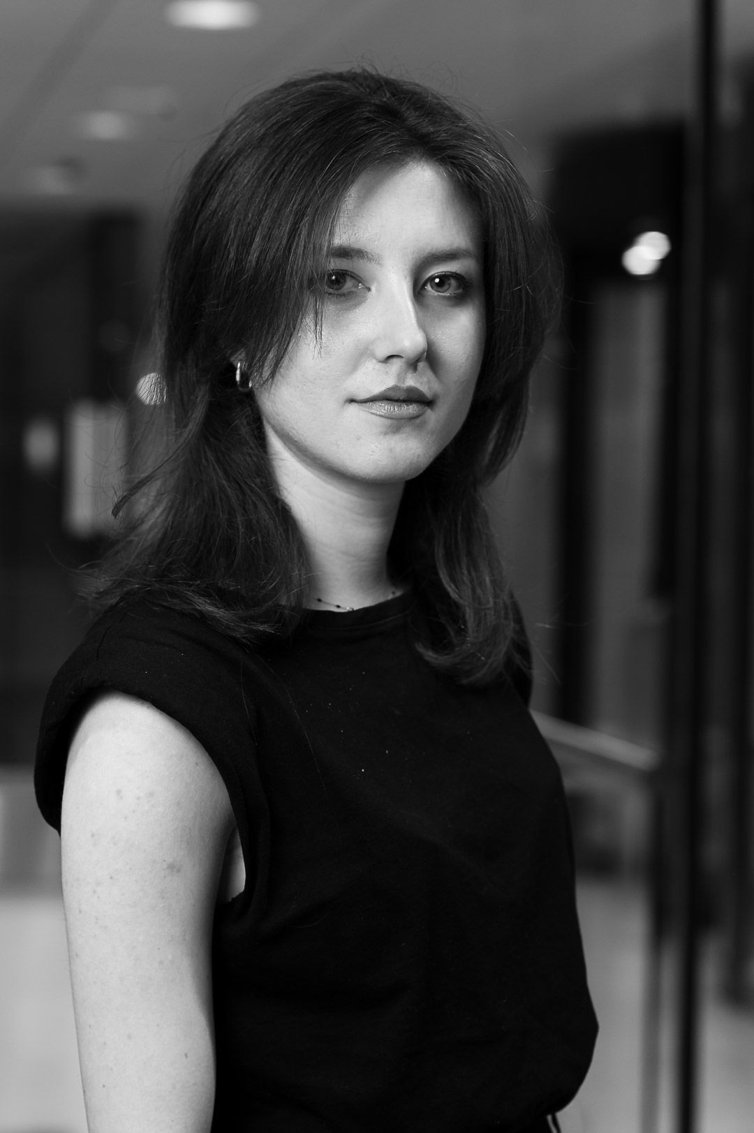 Black and white portrait of someone with long dark hair wearing a sleeveless black top in an indoor hallway setting.