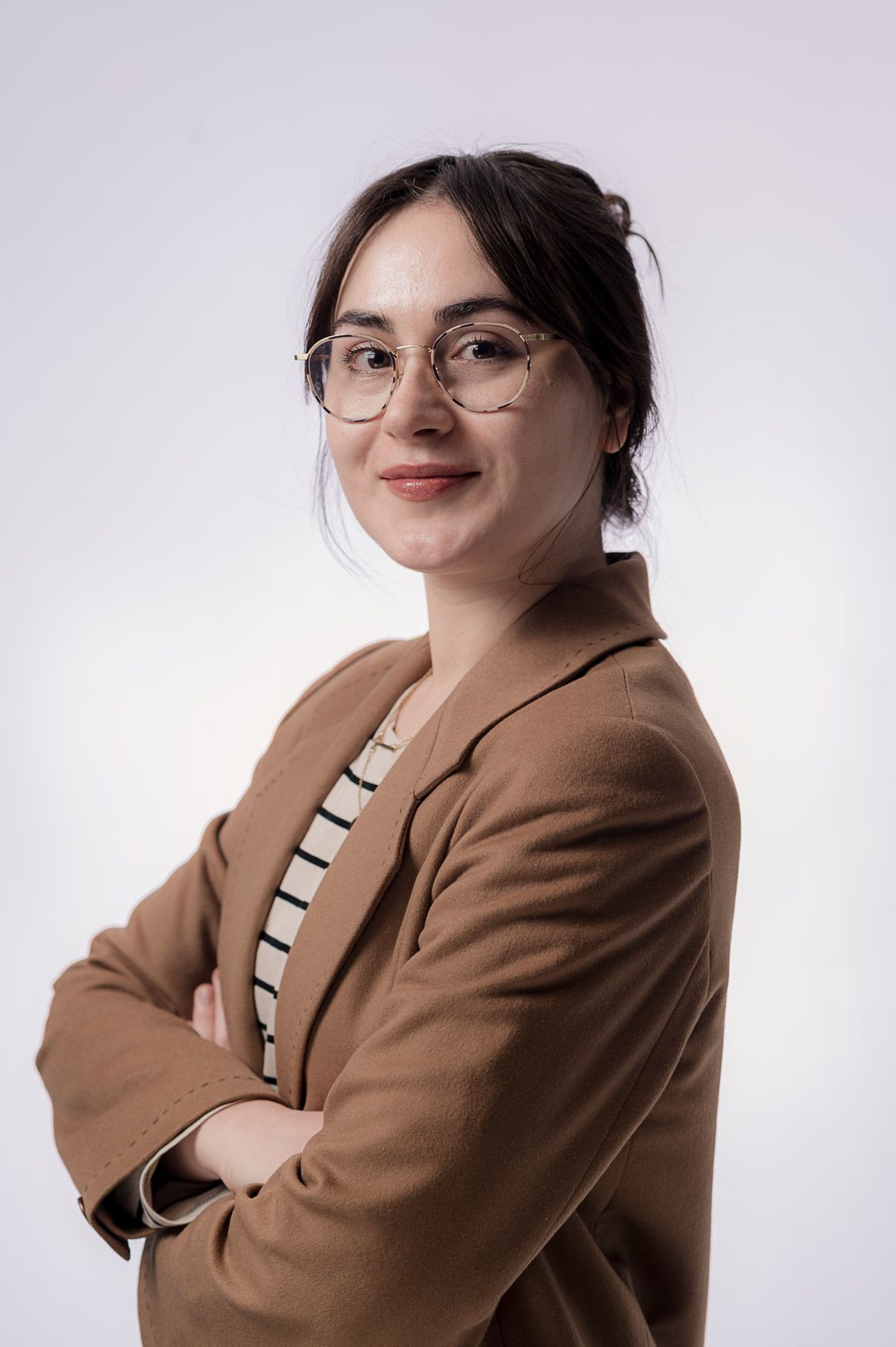 Professional portrait of someone wearing glasses, a brown blazer and striped shirt against a light background.