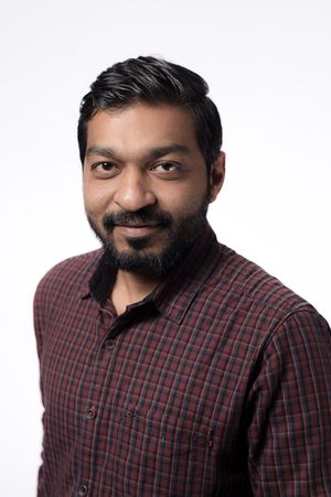 Professional headshot portrait of person wearing a red and black plaid button-down shirt against light background.