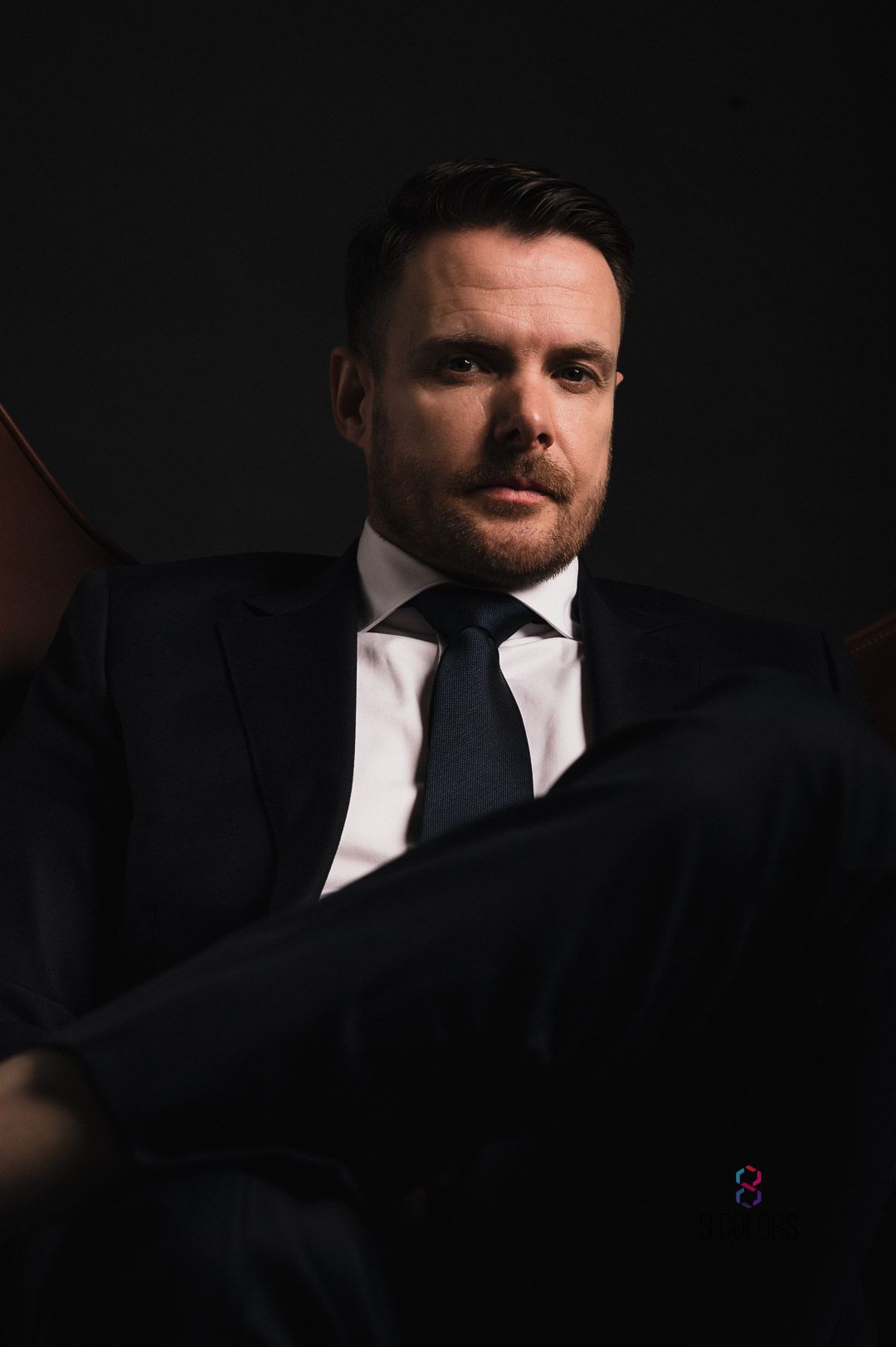 Professional portrait against dark background showing person in black suit and navy tie with dramatic moody lighting.