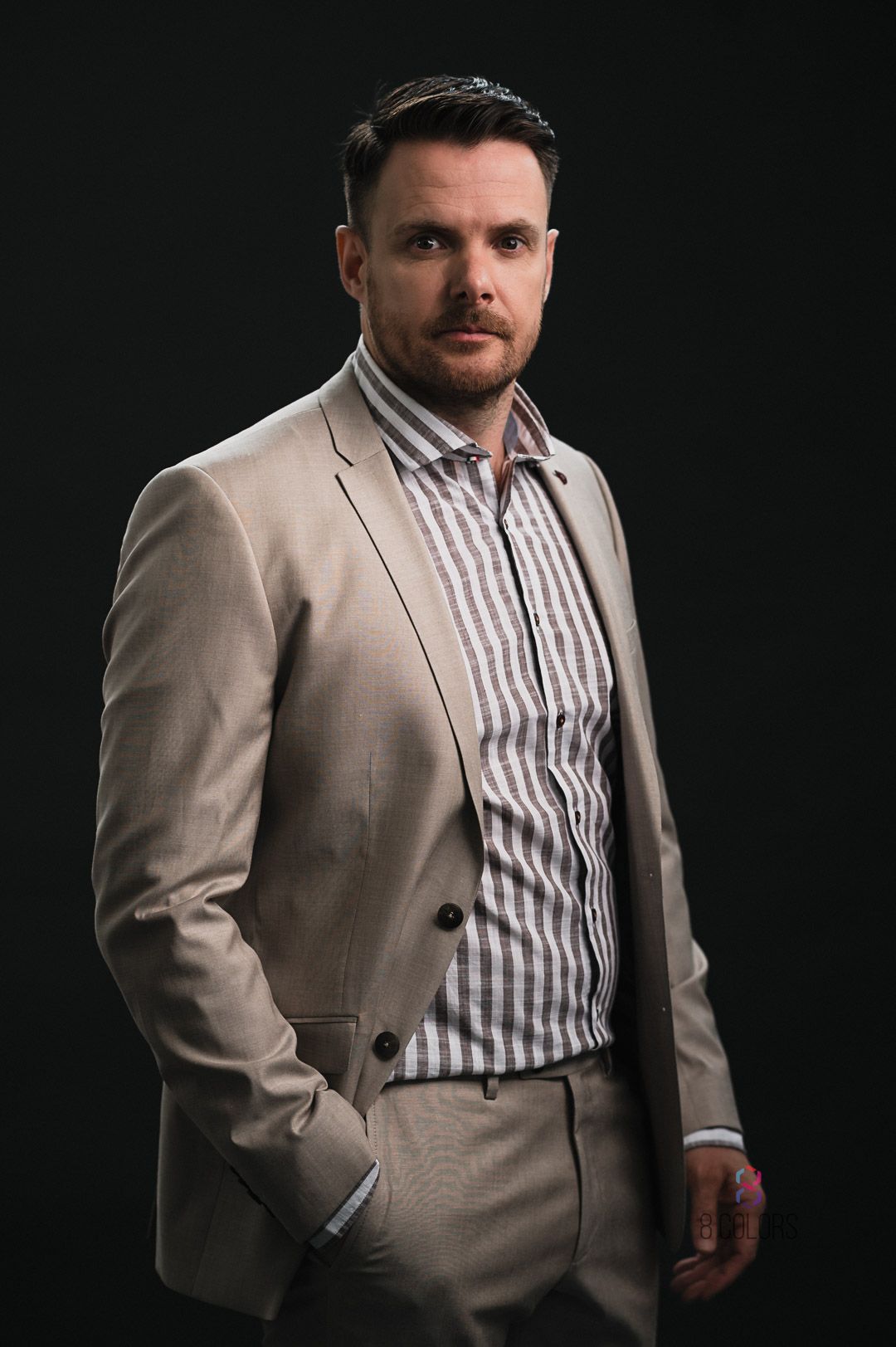 Professional studio portrait of person in beige suit and striped dress shirt against dark background.