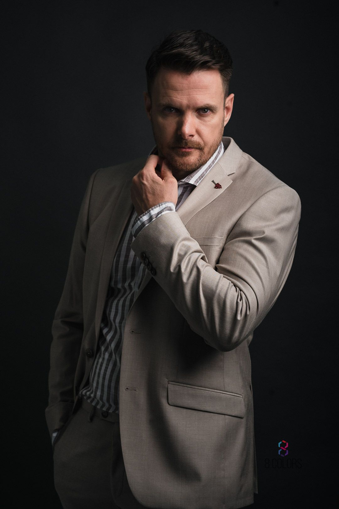 Professional portrait against dark background showing person in beige suit adjusting collar with dramatic lighting.