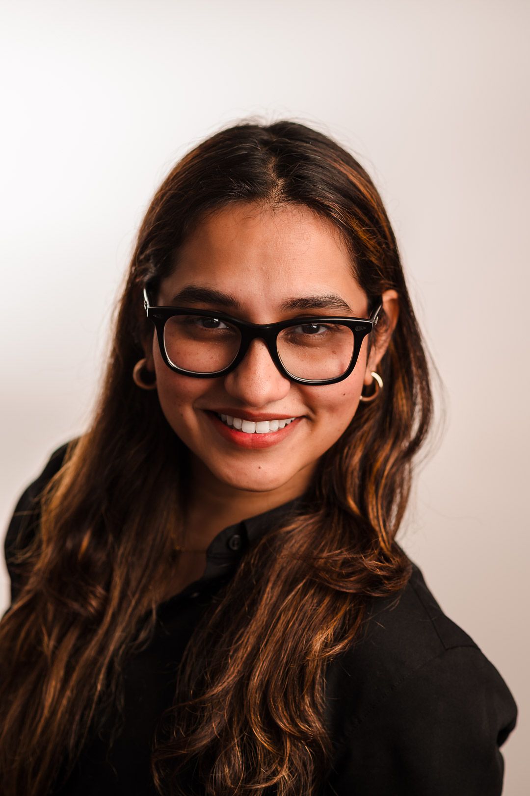 Professional portrait shows person wearing black glasses and dark top, warmly smiling against a light background.