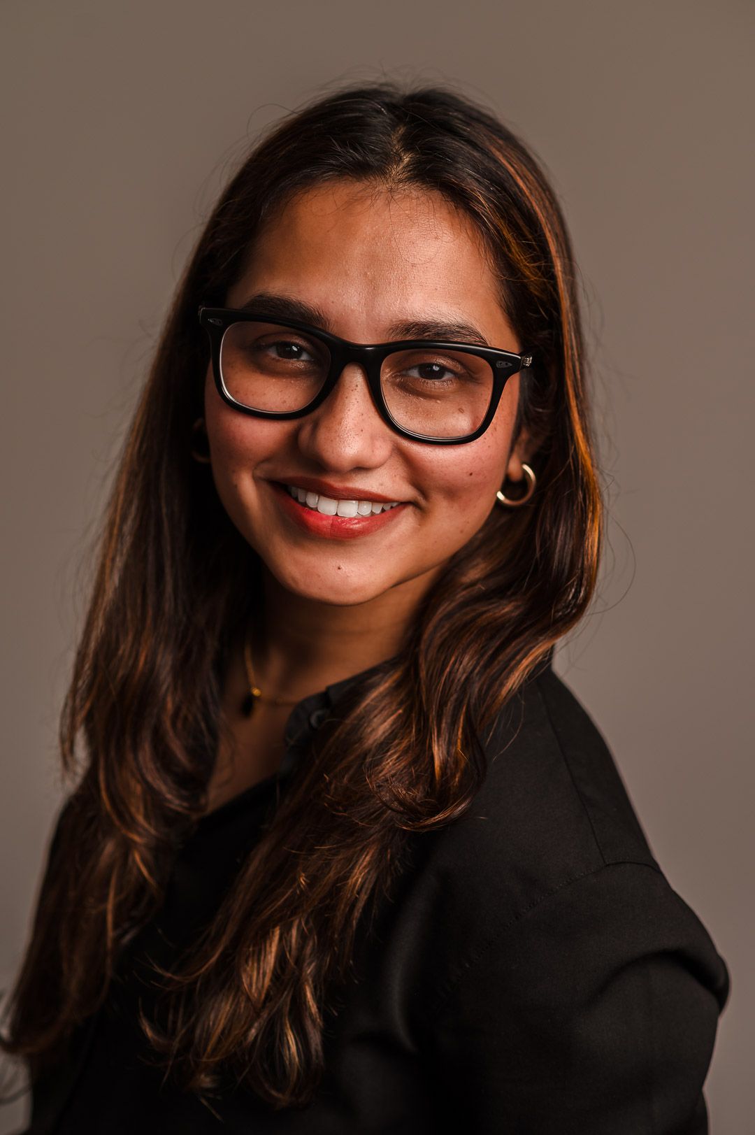 Professional portrait of someone wearing black-rimmed glasses and a dark blazer smiling against a neutral background.