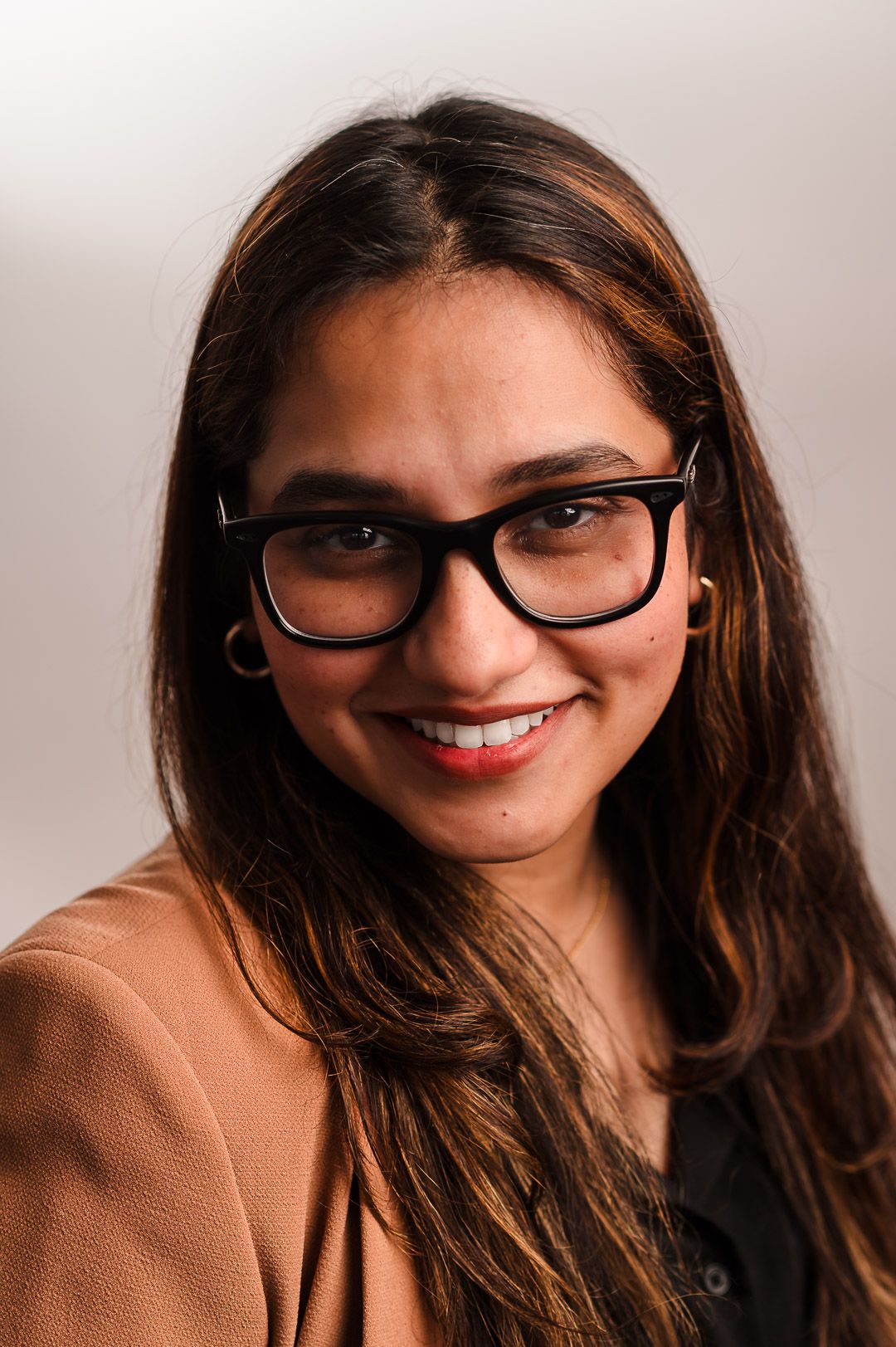 Professional portrait of someone in glasses wearing a tan blazer and black top, smiling warmly at the camera.