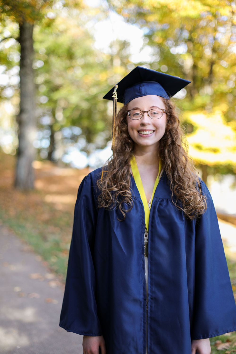 Graduate wearing navy blue cap and gown poses outdoors with autumn foliage in background.