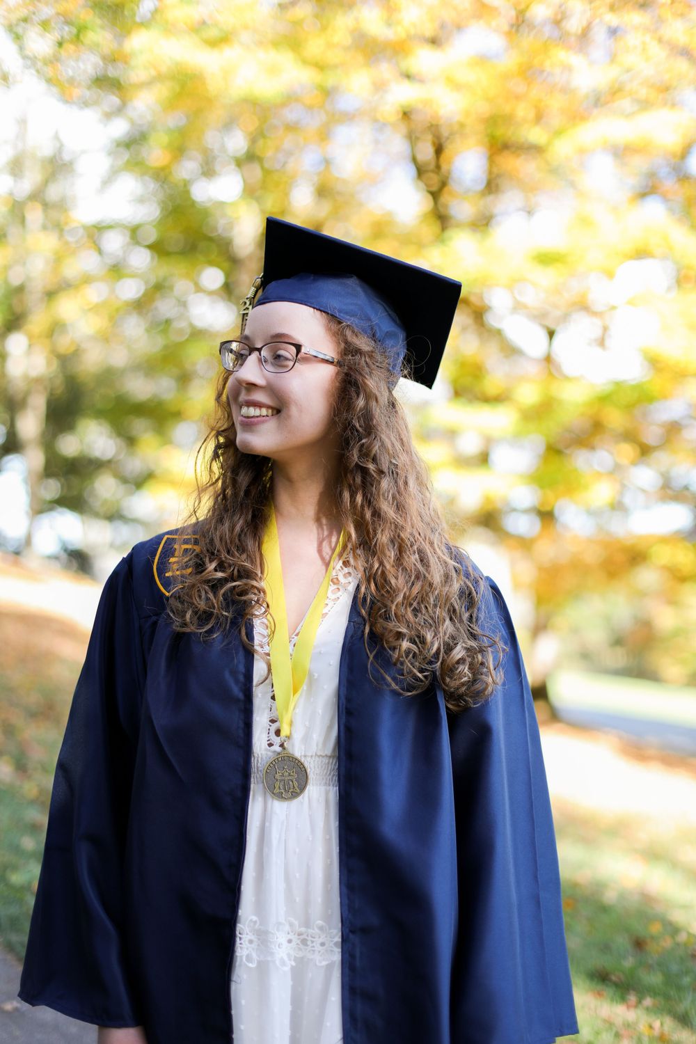 Graduate in navy cap and gown smiles against blurred fall trees.
