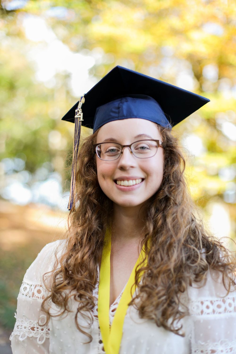 Graduate in white lace dress and navy cap poses with bright autumn scenery.