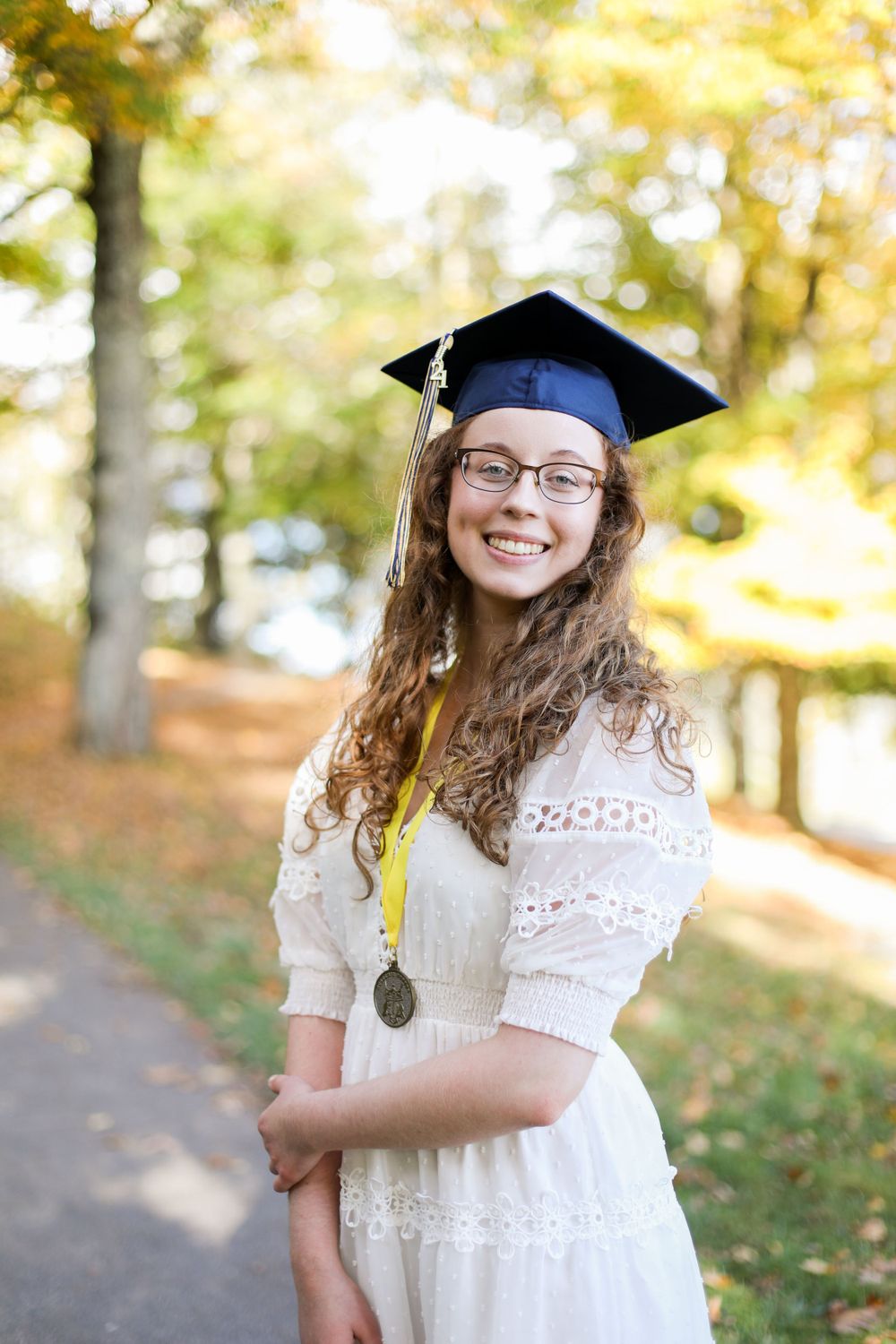 Graduate wearing white eyelet dress and graduation cap poses in autumn setting.