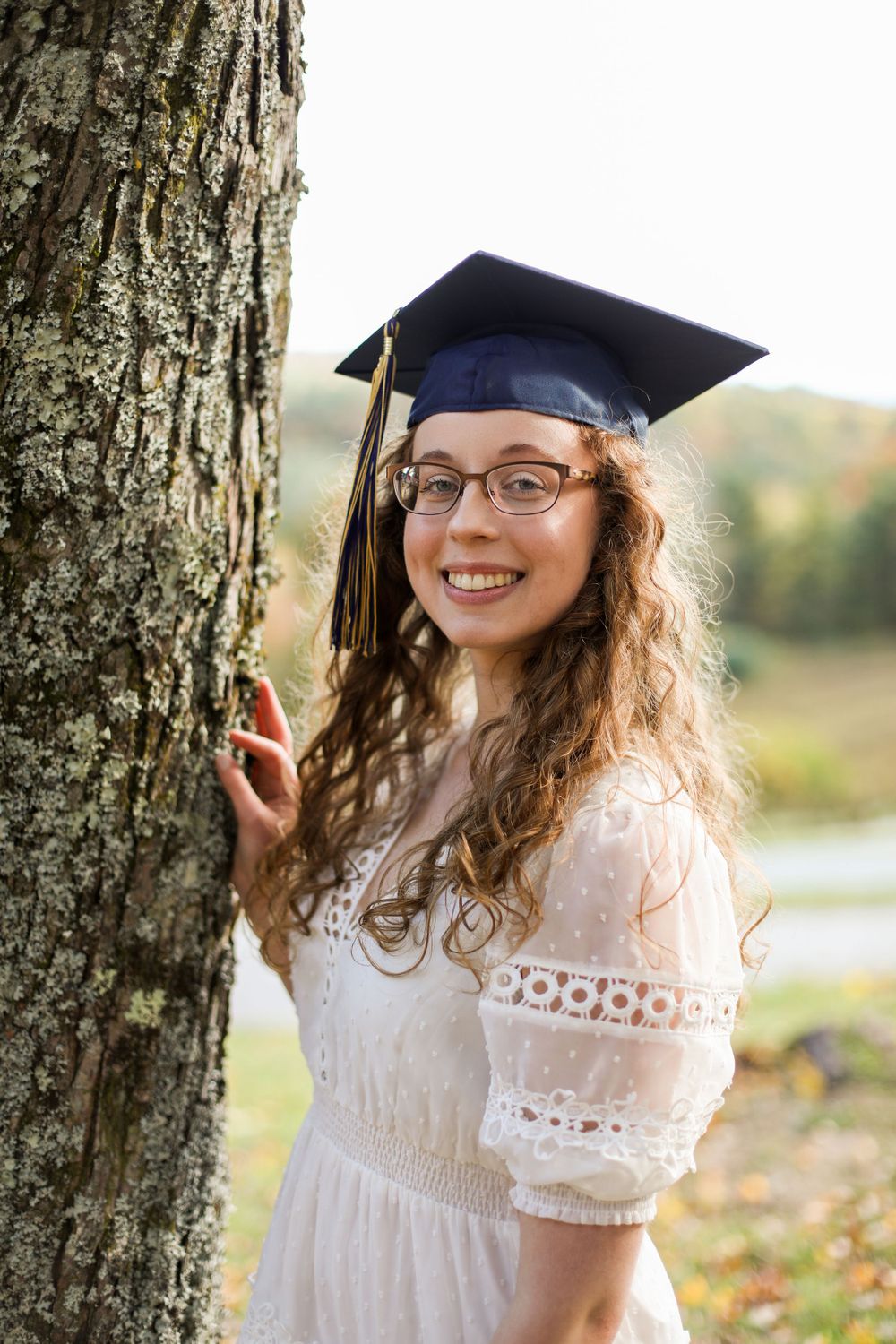 Graduate in white dress poses by tree trunk with fall foliage in background.