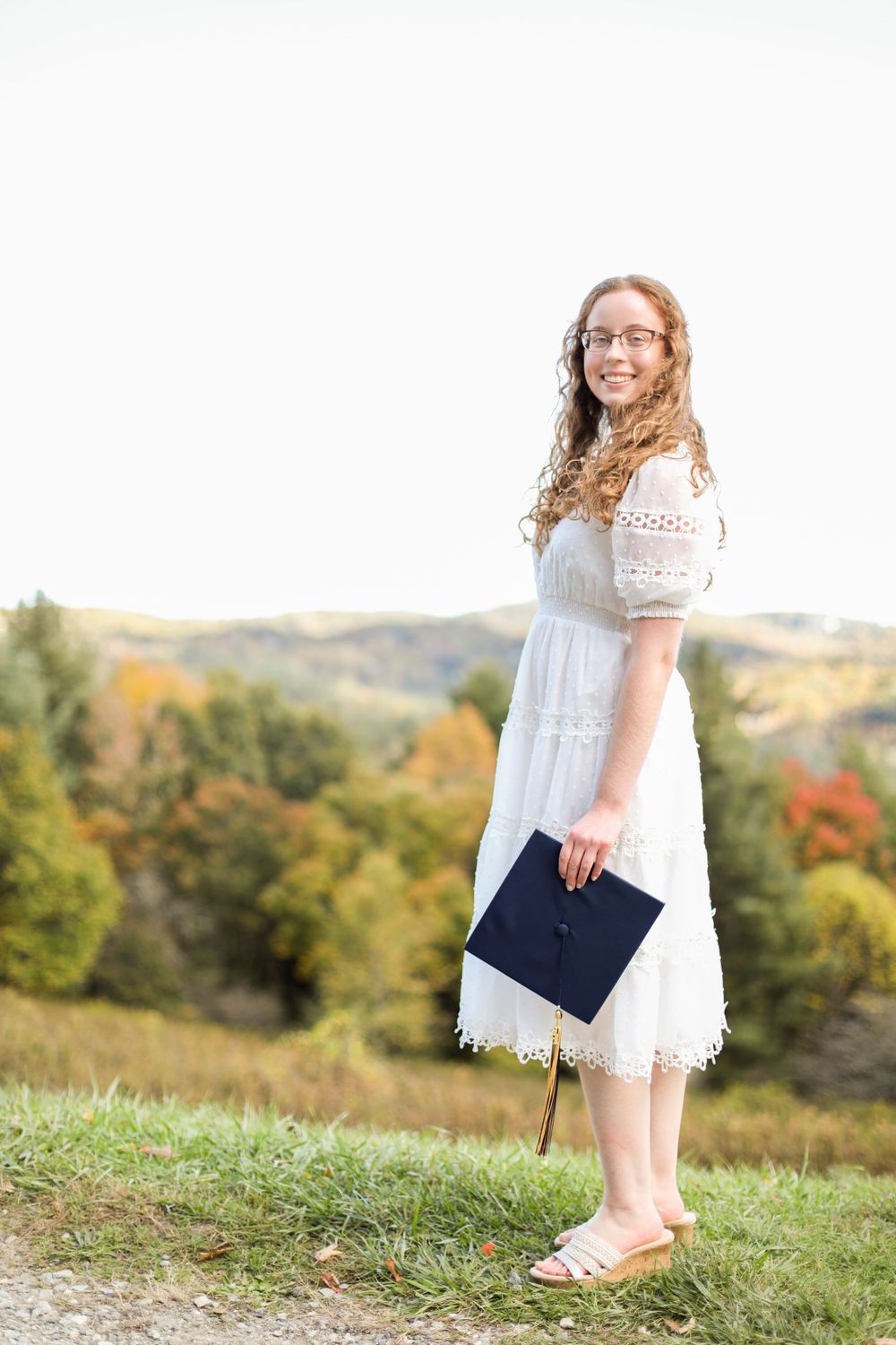 Graduate in white dress holds diploma on grassy hillside with autumn landscape.