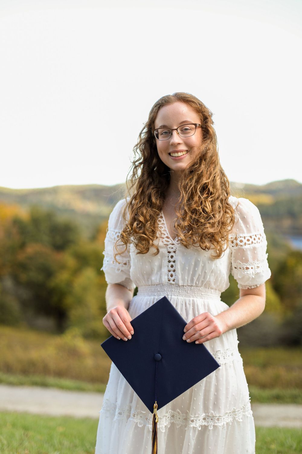 Graduate in white lace dress holds navy blue diploma cover outdoors.