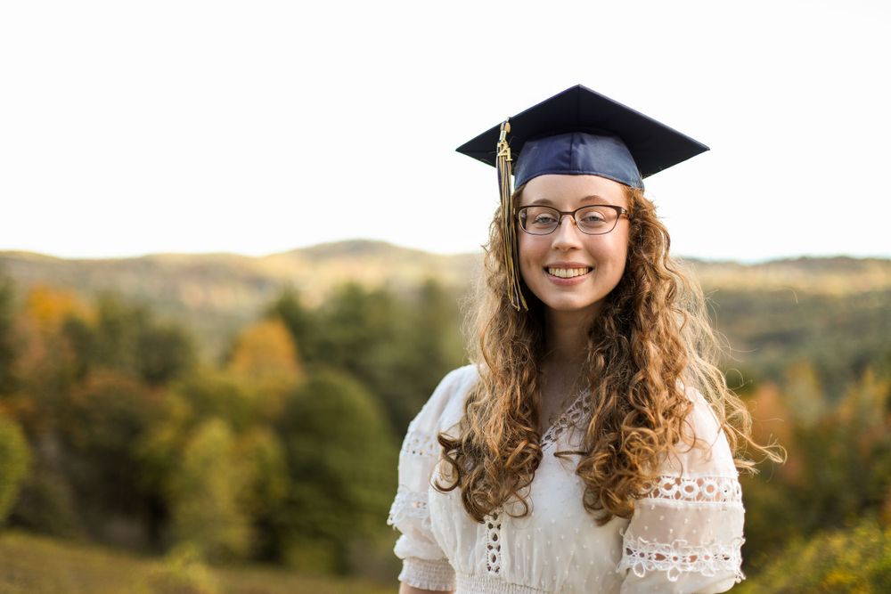 Graduate poses against scenic autumn hillside in white dress and cap.