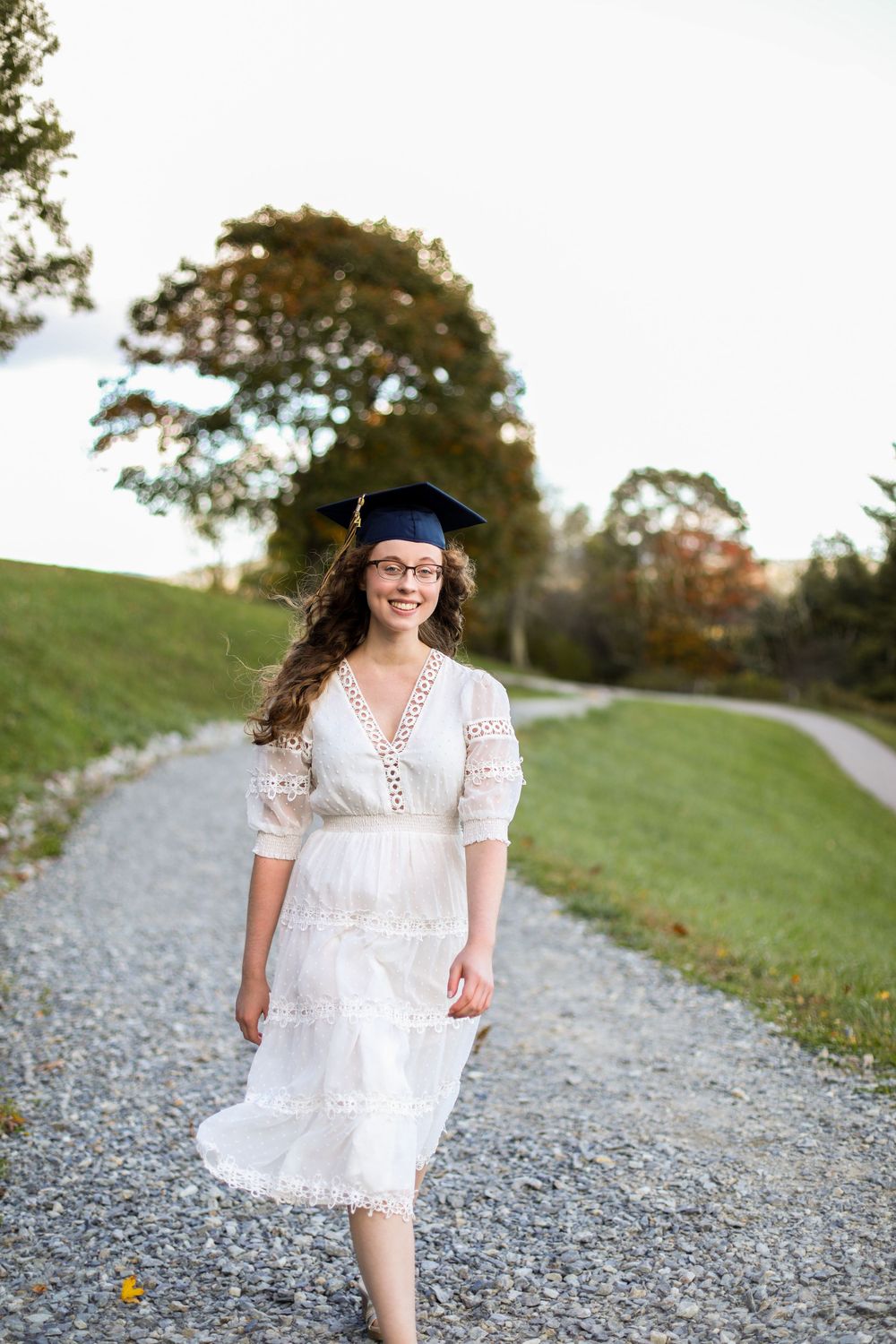 Graduate walks along gravel path in white dress and navy graduation cap.