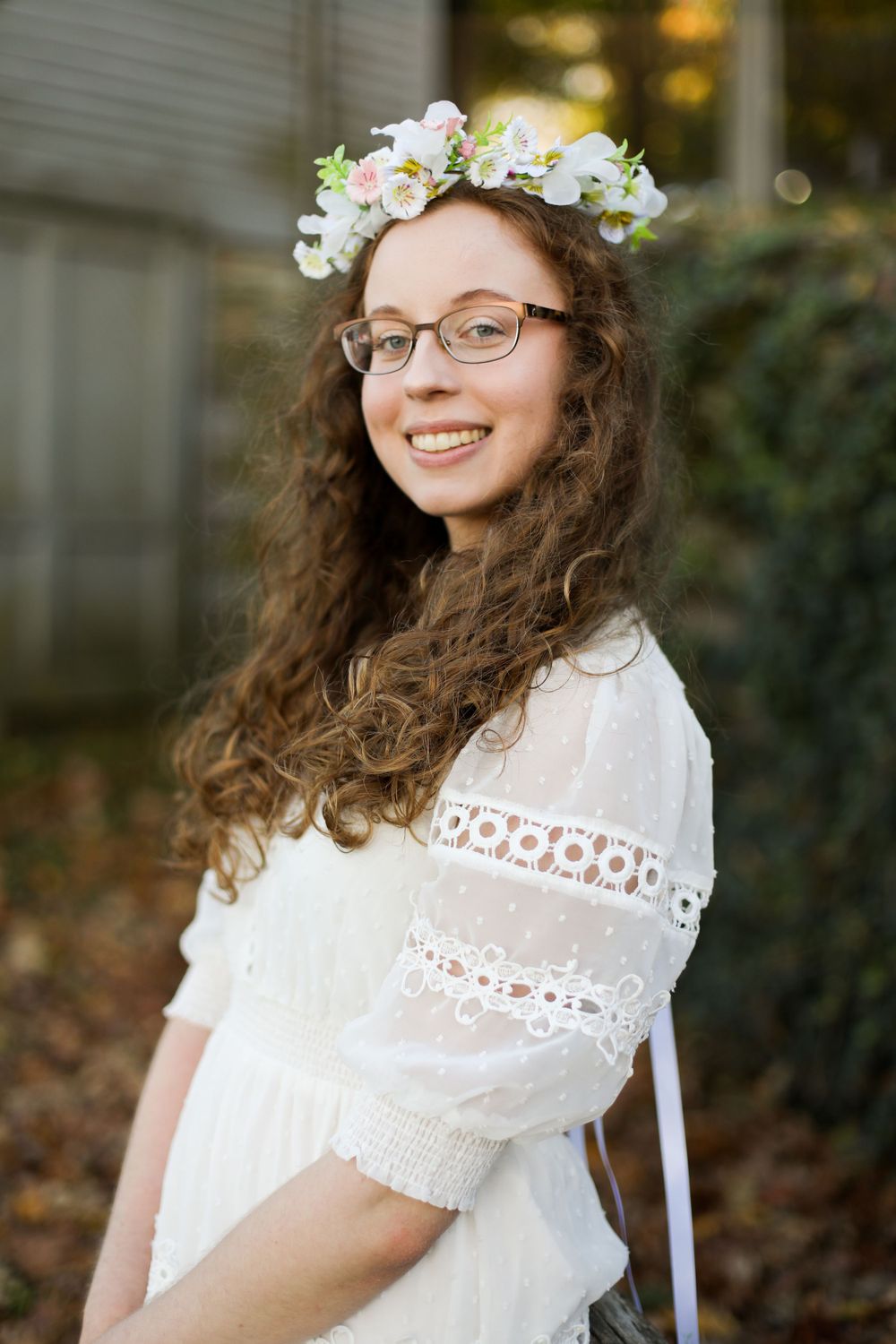 Graduate wearing floral crown and white lace dress smiles in outdoor setting.