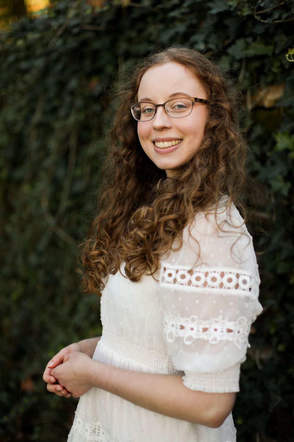 Graduate wearing a white lace dress smiles while posing for portraits in a garden setting.