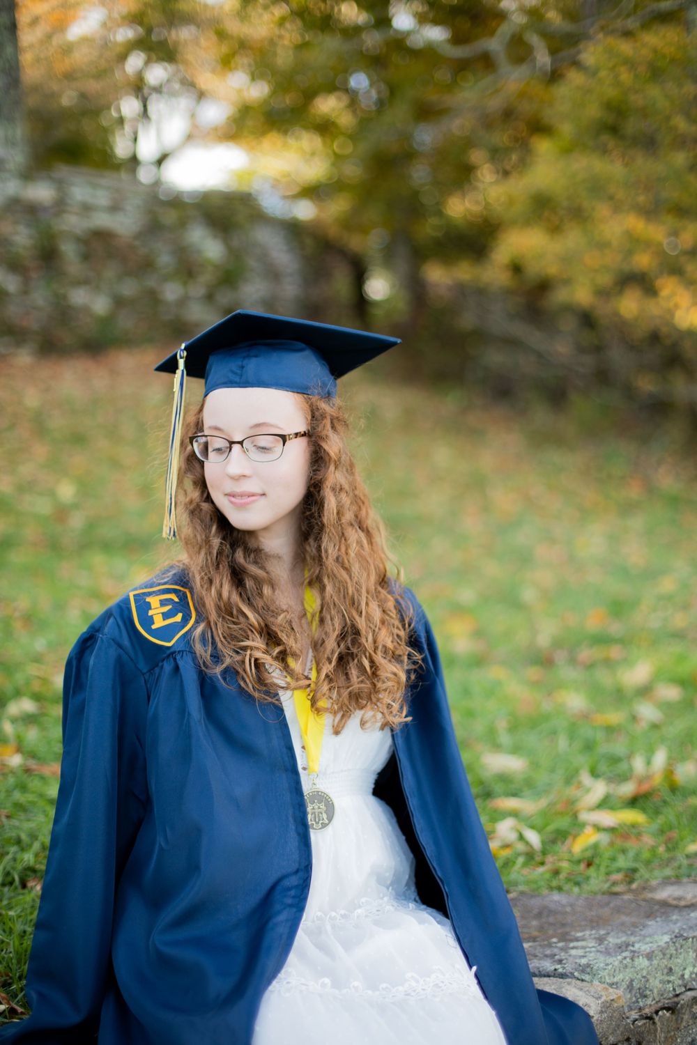Graduate in blue cap and gown poses outdoors during a fall photoshoot.