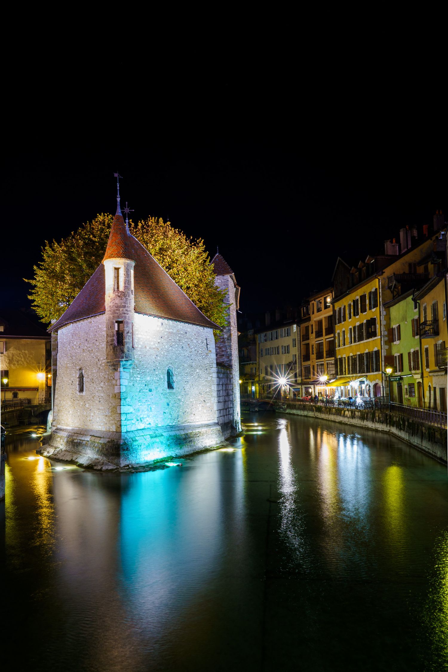 Historic stone chapel illuminated in blue light reflects in calm canal waters at night.