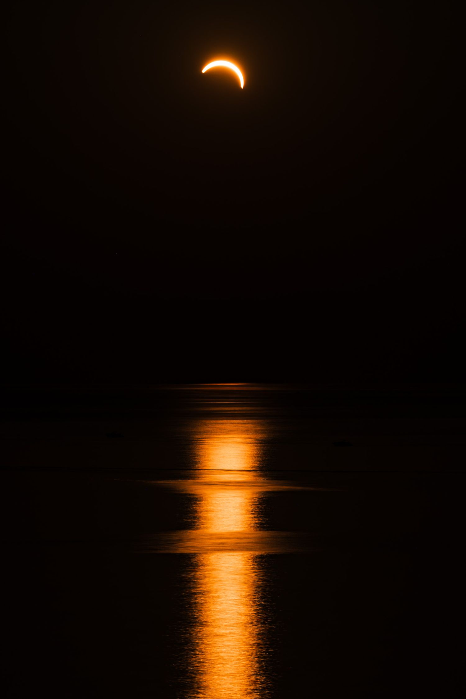 Partial solar eclipse over dark ocean waters with vivid orange reflection glowing on the calm sea surface at night.