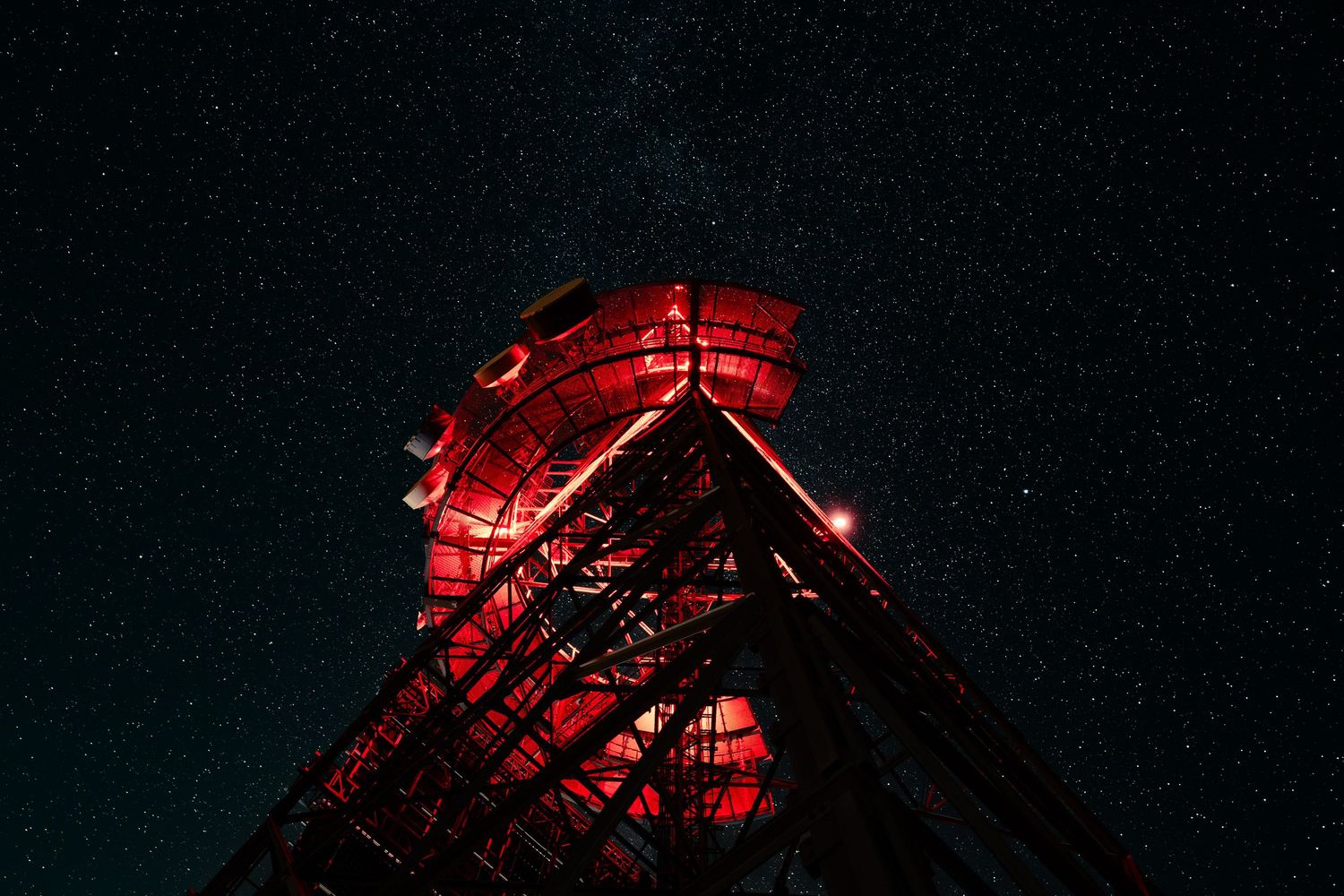 Red illuminated communication tower against a stunning starry night sky, viewed from below at a dramatic angle.