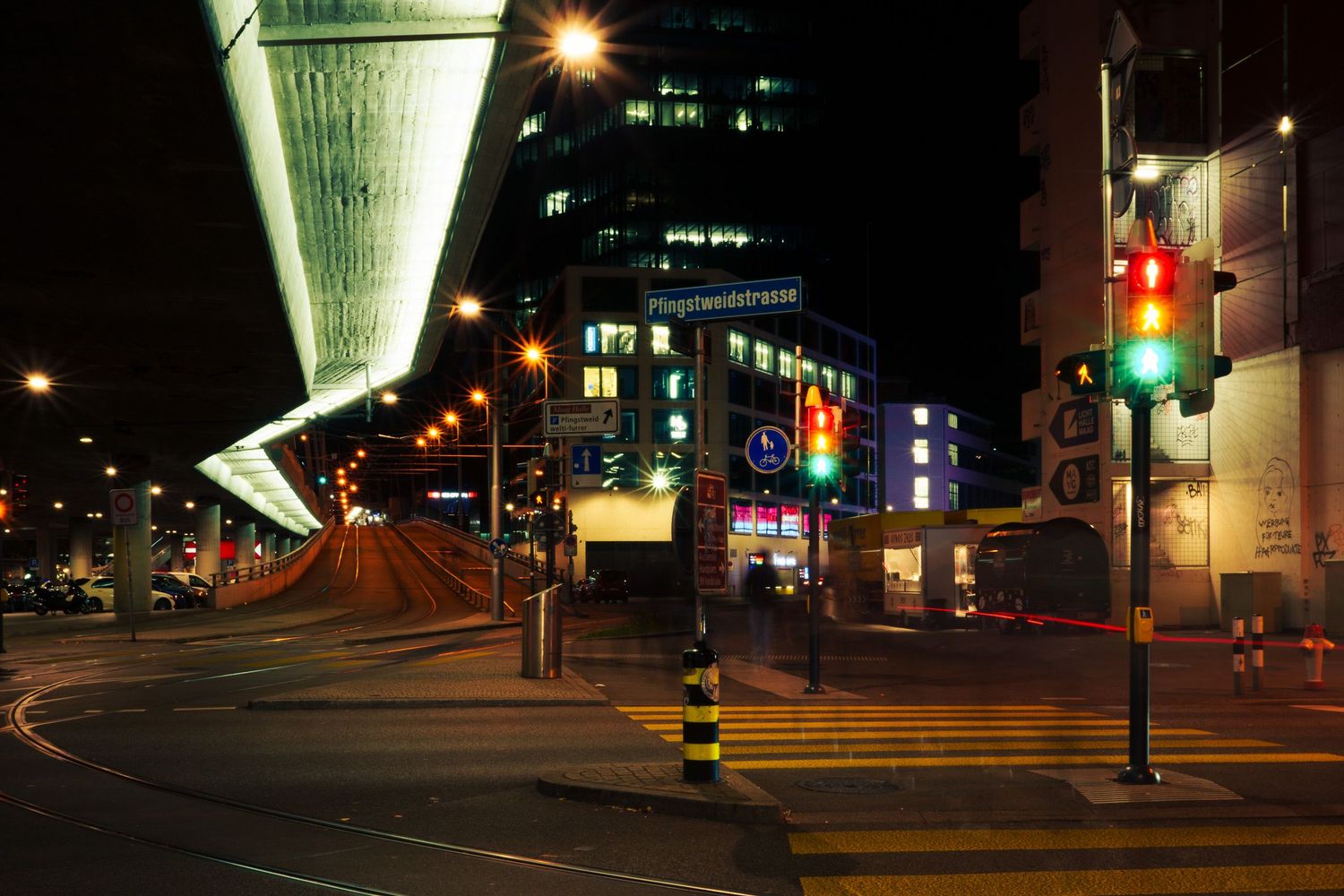 Nighttime city intersection with glowing traffic lights, illuminated buildings, and a modern elevated structure in an urban setting.