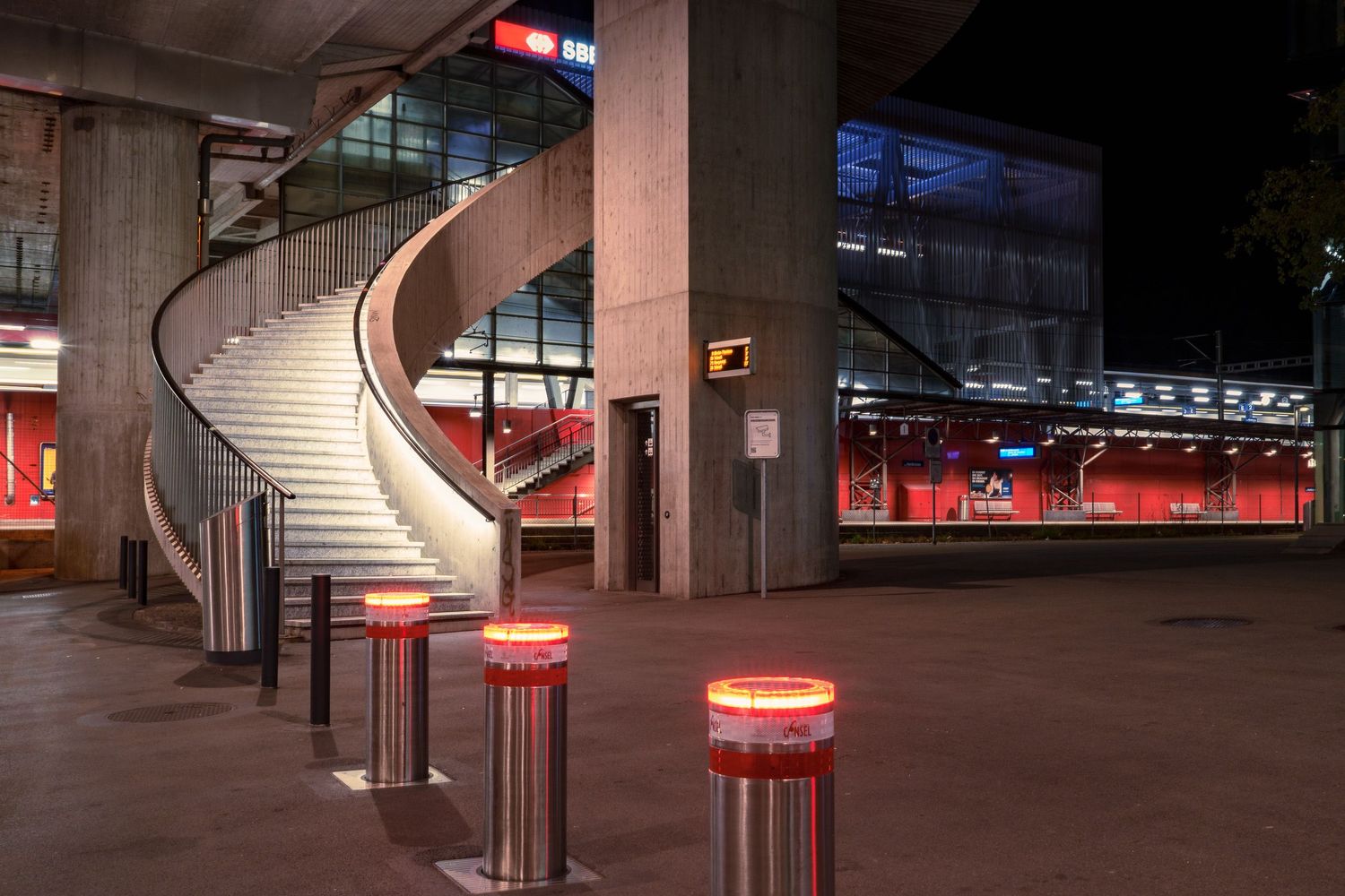 Illuminated bollards line the entrance of a modern stadium at night, featuring a dramatic curved staircase and red seating.