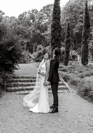 An elegant black and white portrait of a wedding couple standing by stone steps in a garden setting.