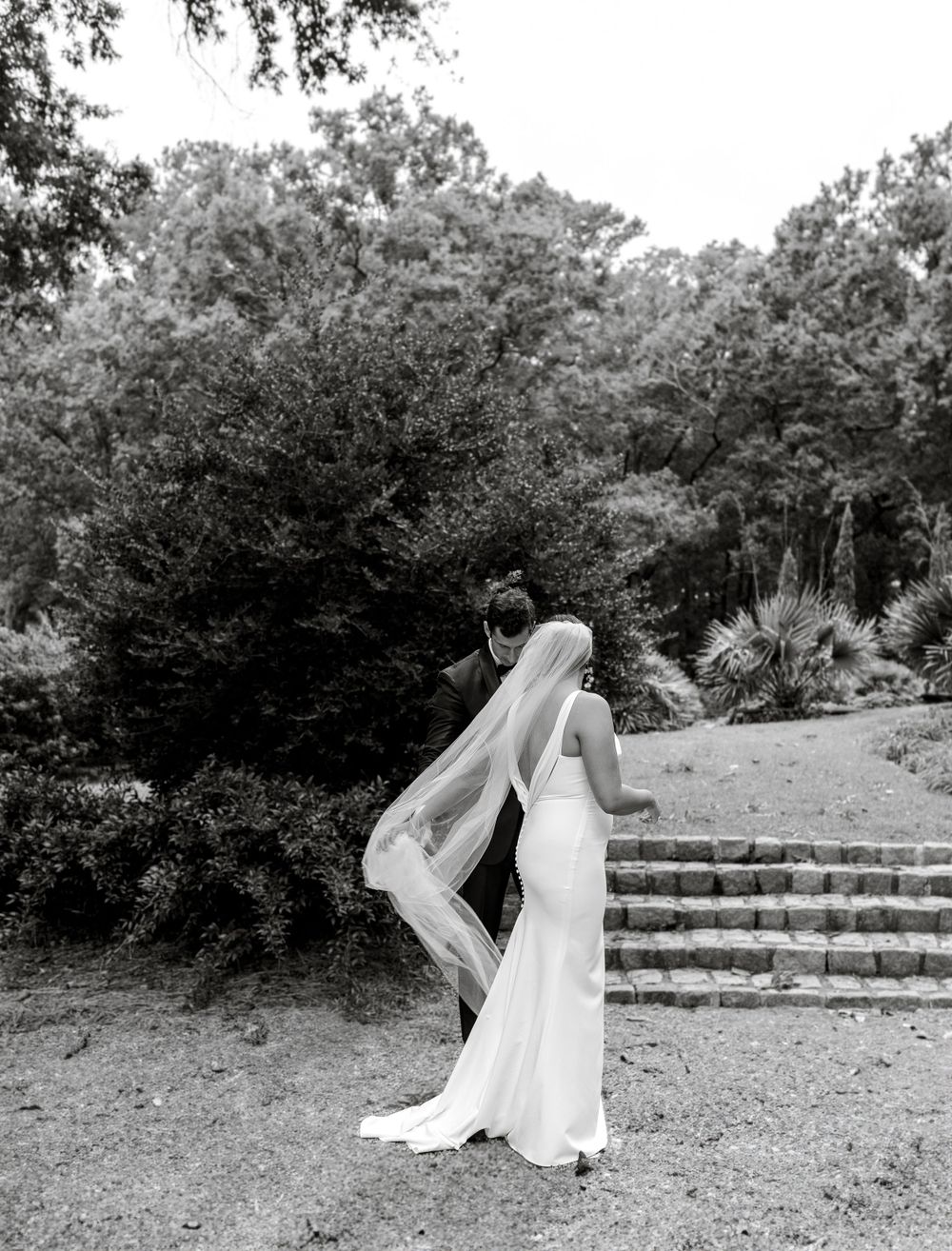 A bride in a flowing white dress stands in a garden setting with stone steps and lush greenery in black and white.