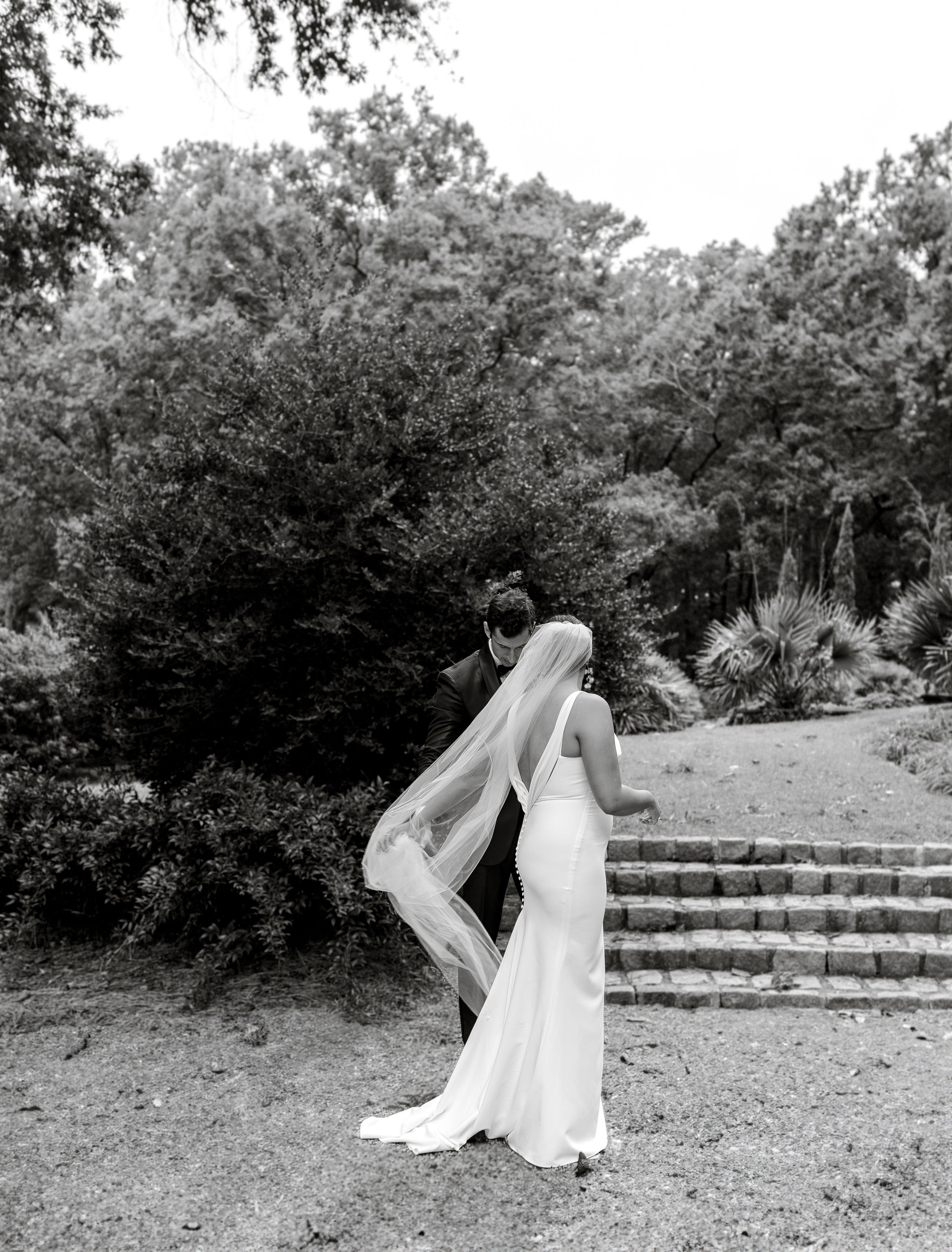 A bride in a flowing white dress stands in a garden setting with stone steps and lush greenery in black and white.