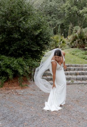 A flowing wedding veil catches the breeze as a bride poses on stone steps surrounded by gardens.