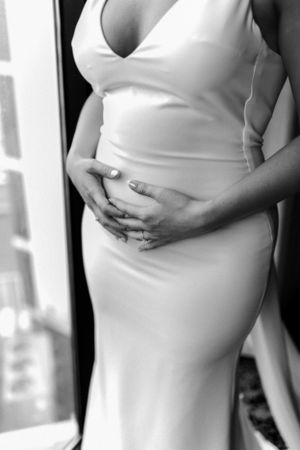 Black and white close-up photo of a pregnant person in a white dress holding their baby bump.