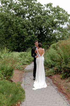 A couple in wedding attire walks down a garden path surrounded by lush greenery.
