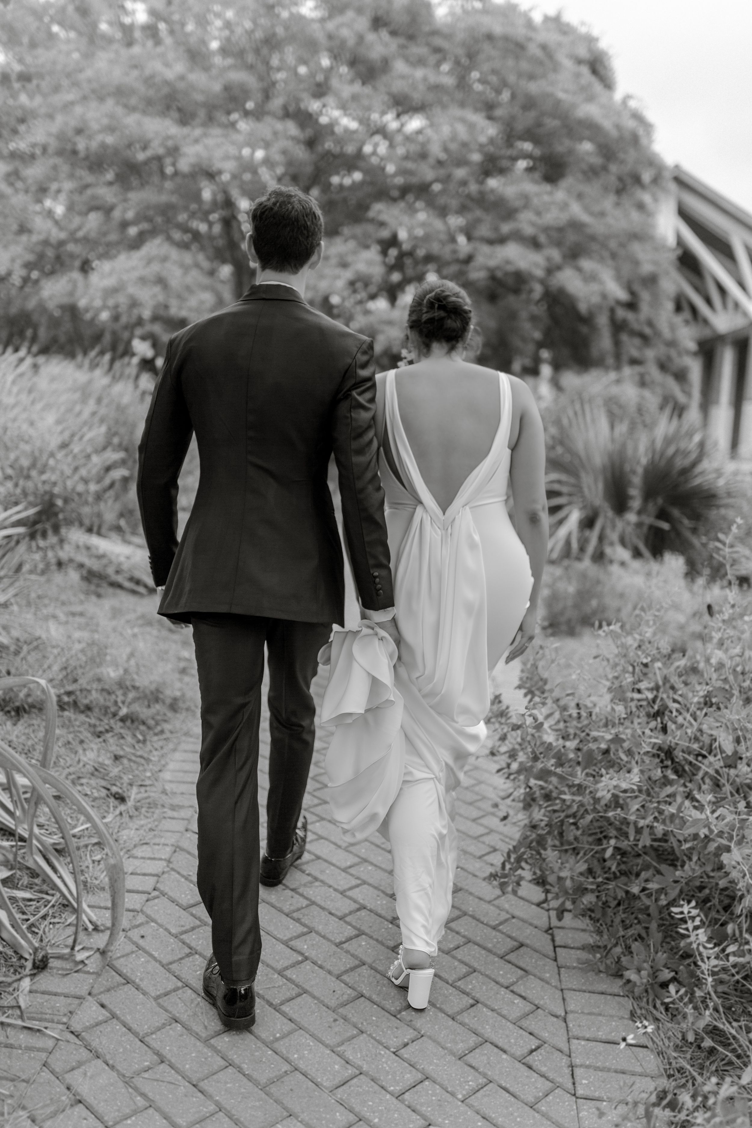 Back view of newlyweds walking together on a brick pathway through a garden setting.