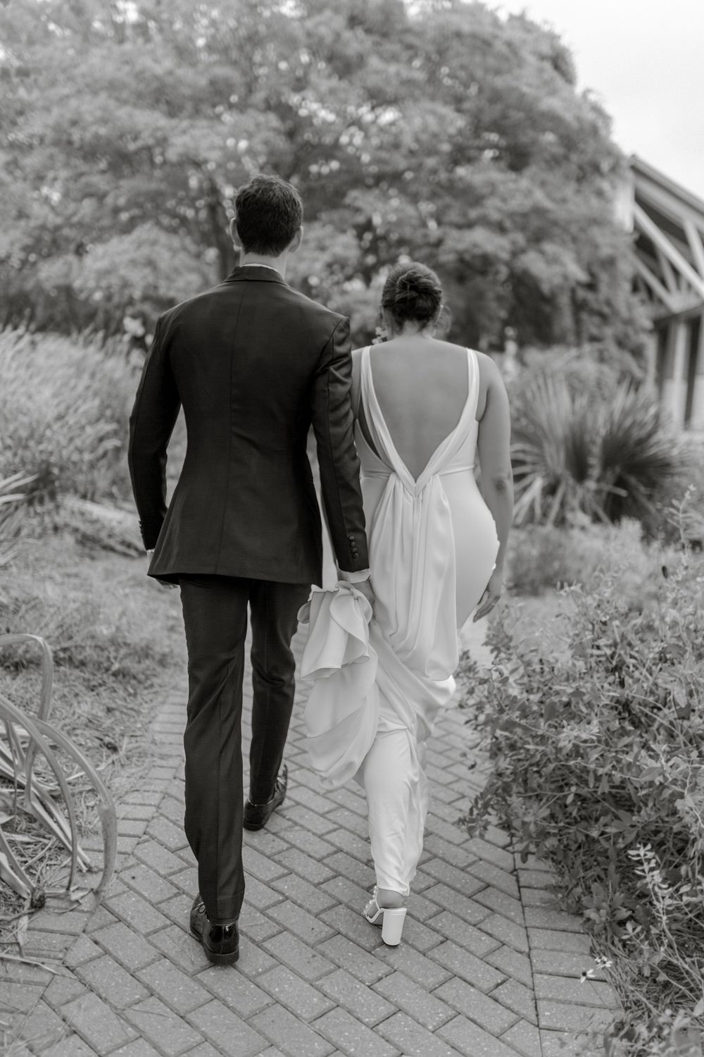 Back view of newlyweds walking together on a brick pathway through a garden setting.