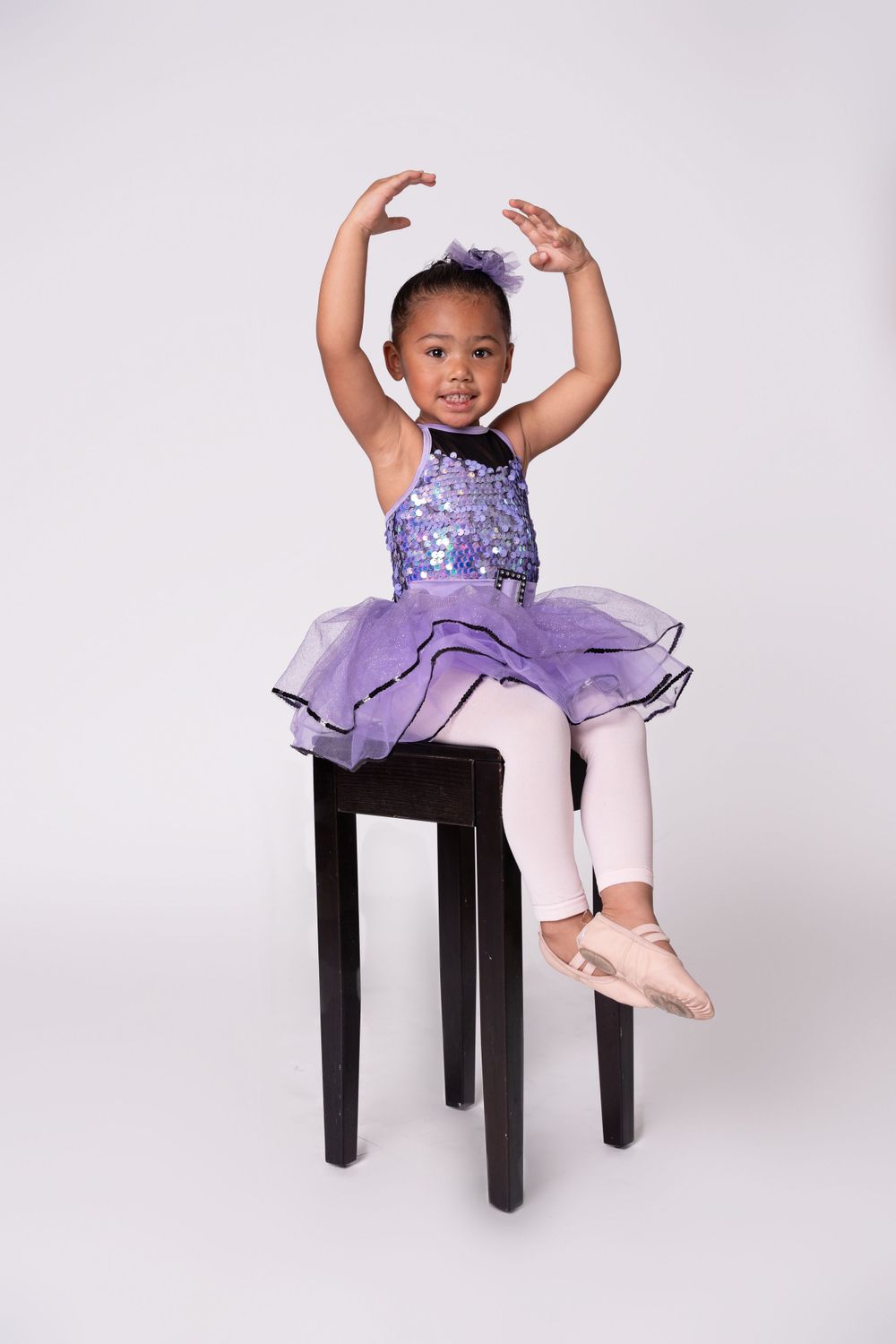Young dancer in purple sequined tutu dress posing gracefully on black stool.