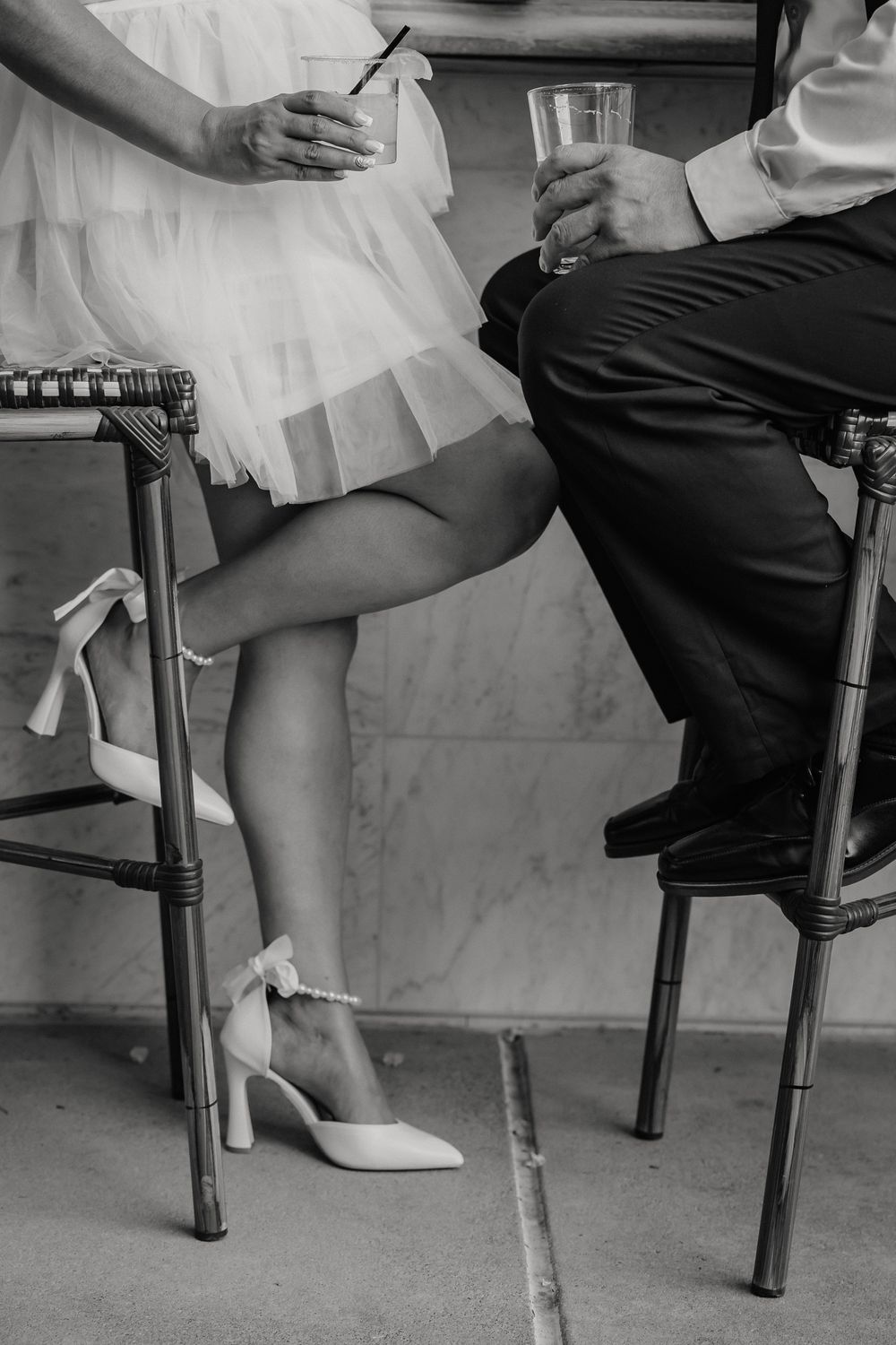 Black and white photo showing couple's legs at bar stools with formal attire and white heels.