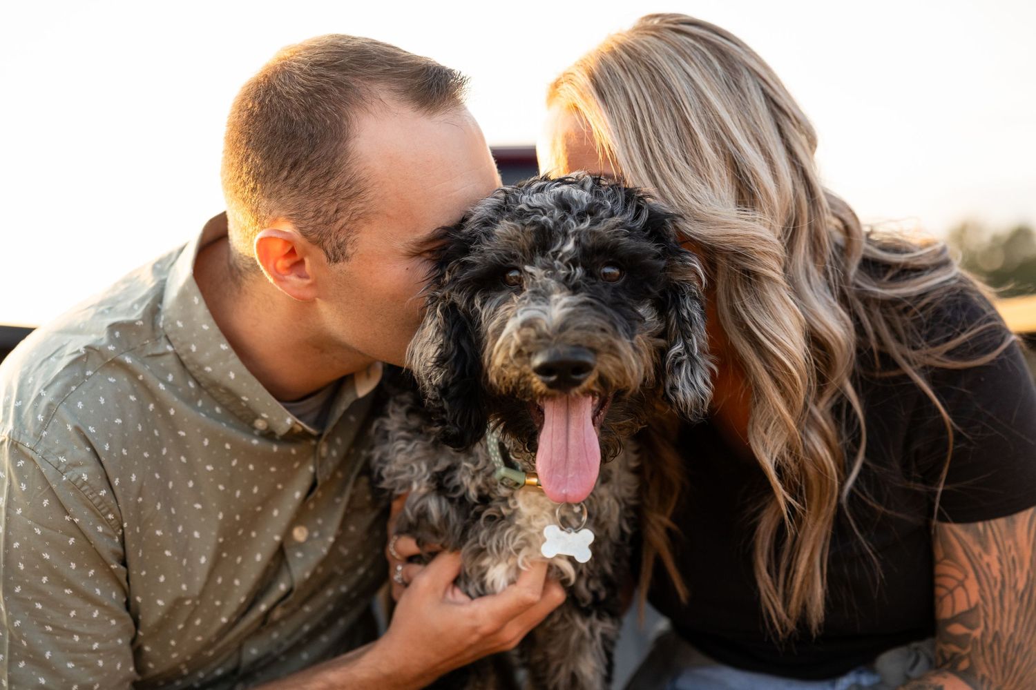 Couple kissing their fluffy black doodle dog outdoors at golden hour.