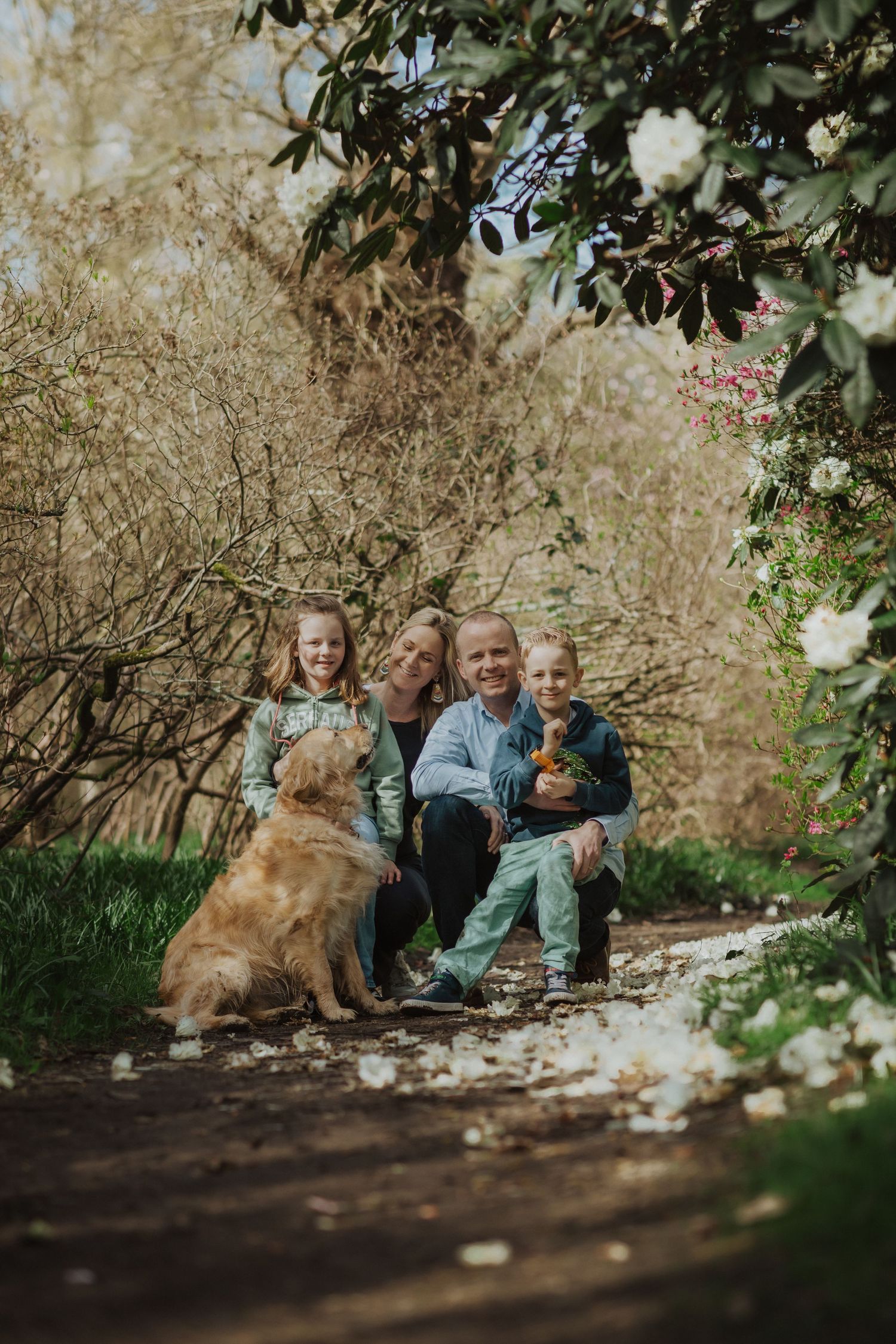 A group sits together on a garden path with their golden retriever under blooming spring flowers.