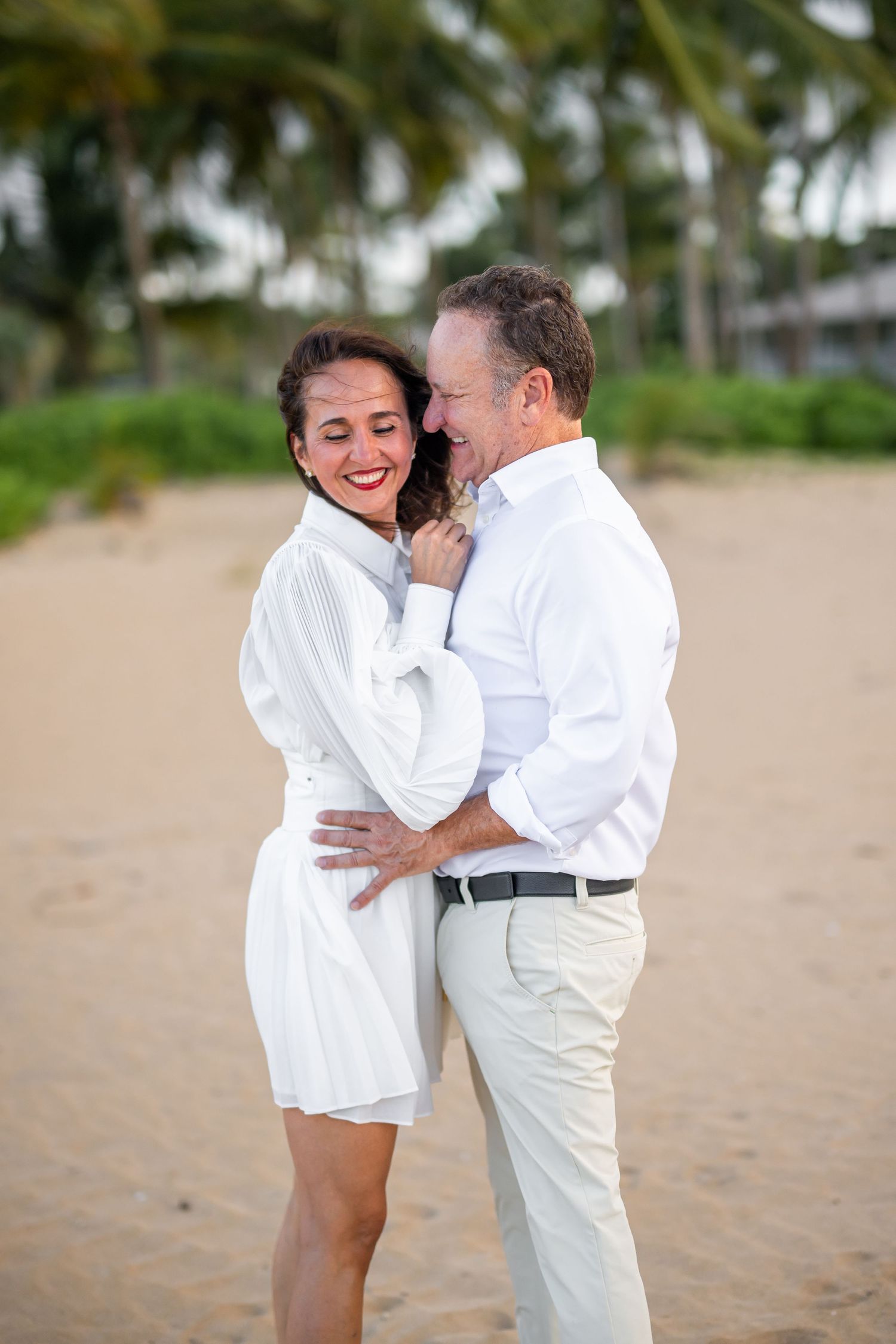 Happy couple in white outfits embracing on a tropical beach with palm trees.
