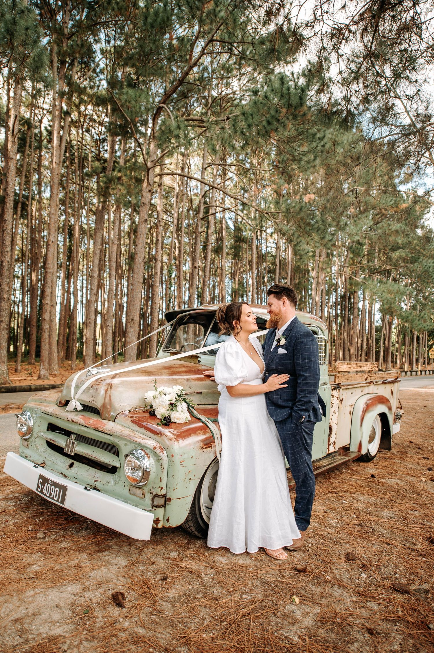 A romantic wedding photo featuring a vintage mint green pickup truck in a pine forest setting.