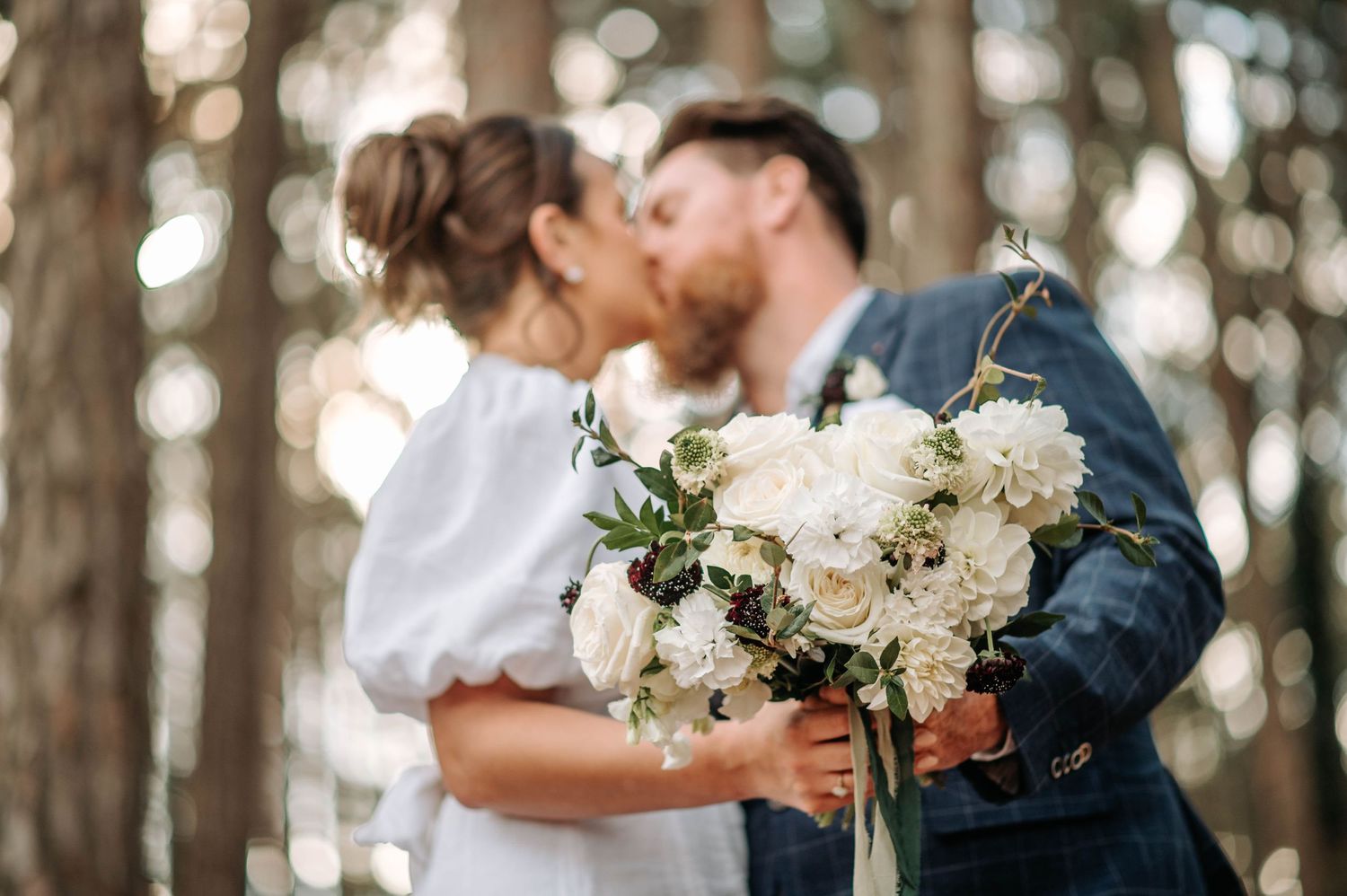 Wedding couple shares kiss while holding elegant white floral bouquet in forest.