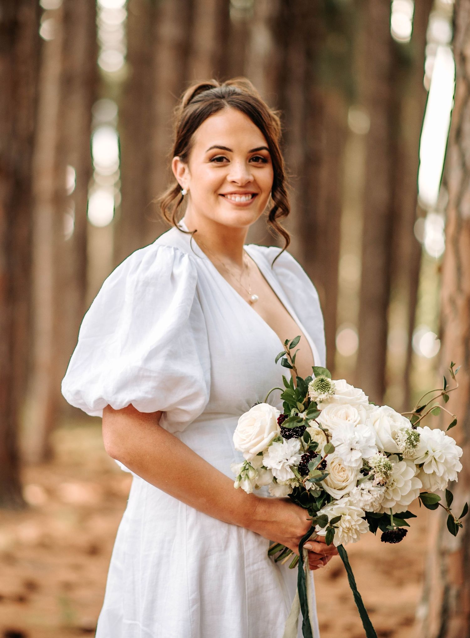 A bride in a white dress holds a bouquet of white flowers against a forest backdrop.