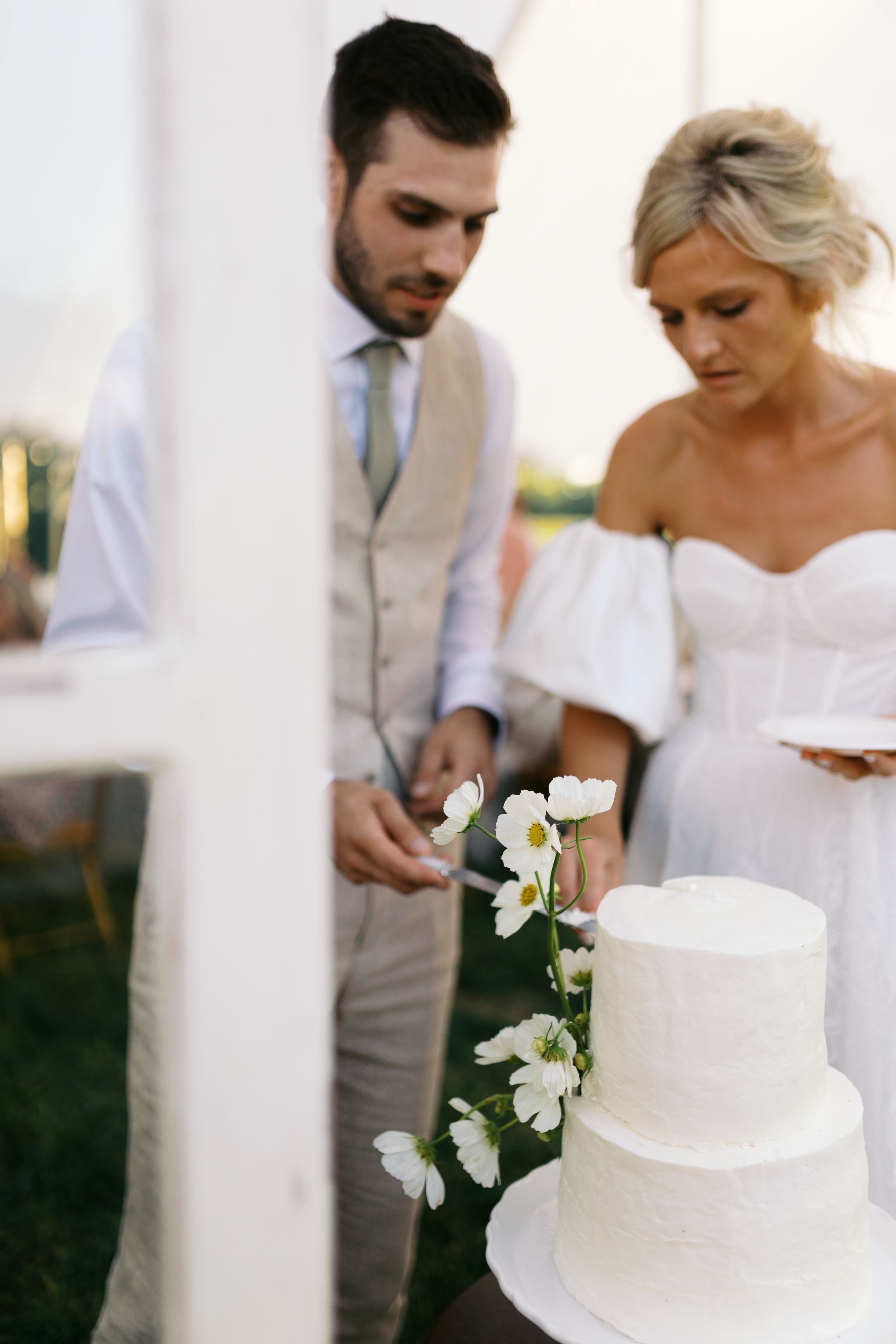Bride and groom cutting a two-tier white wedding cake adorned with white flowers at an outdoor wedding reception.