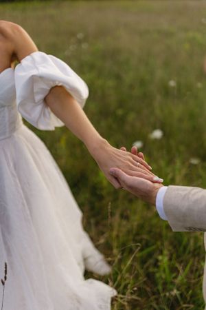Close-up of newlyweds holding hands in a sunlit meadow with flowing white dress.