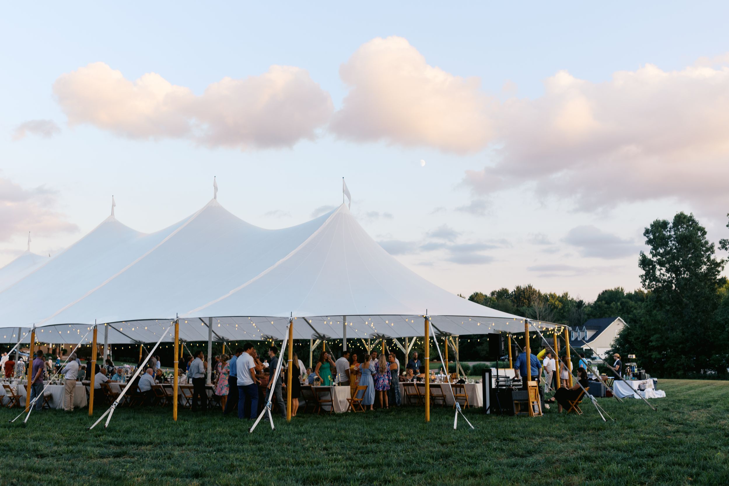 Elegant white sailcloth wedding tent on green grass field at dusk with guests gathered underneath.