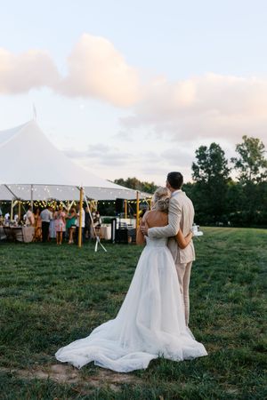 Bride and groom embracing outdoors at sunset near a white tent wedding reception with string lights on green grass.