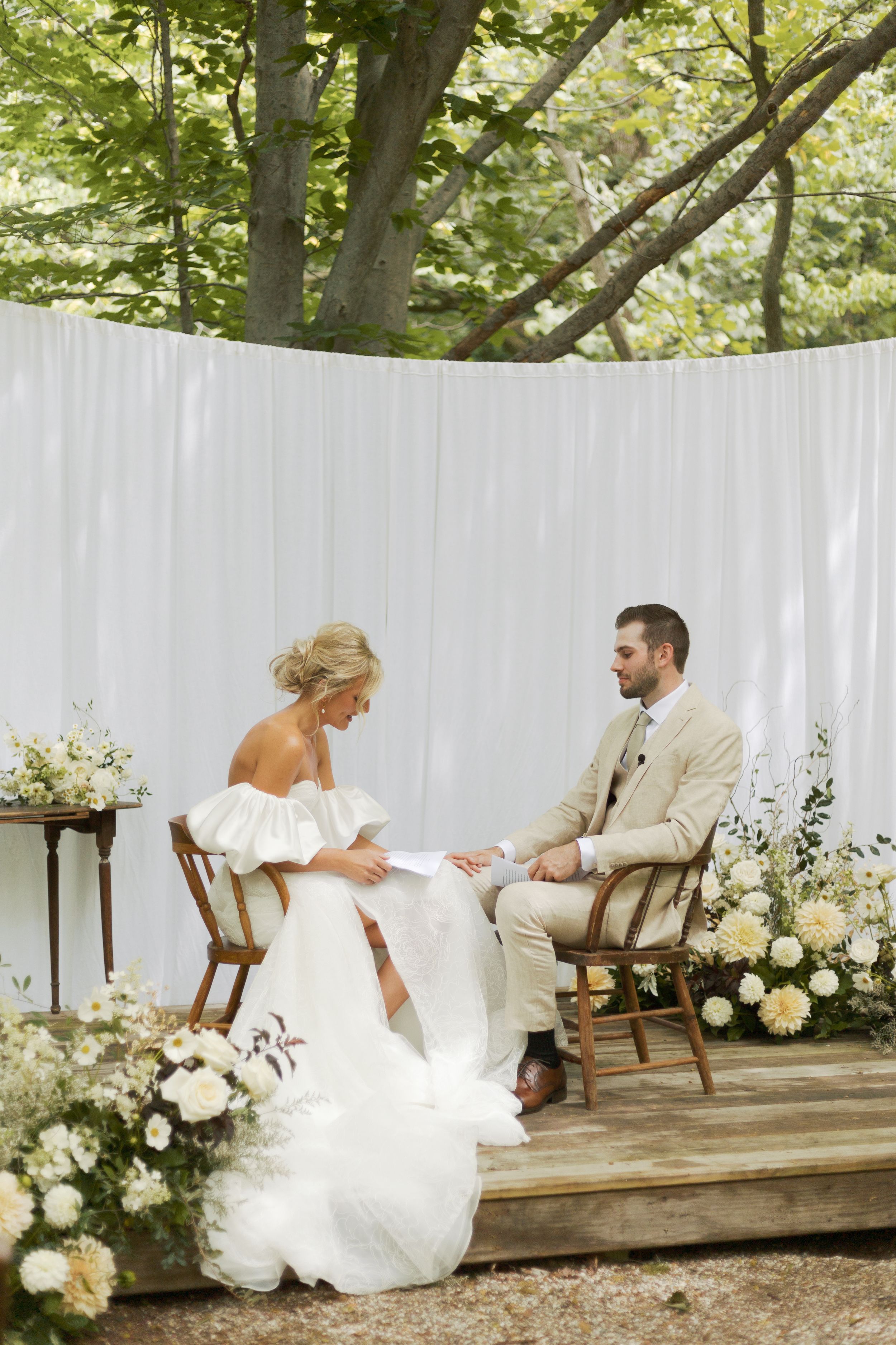 Bride and groom exchange vows seated on wooden chairs at an outdoor wedding ceremony surrounded by white floral arrangements.