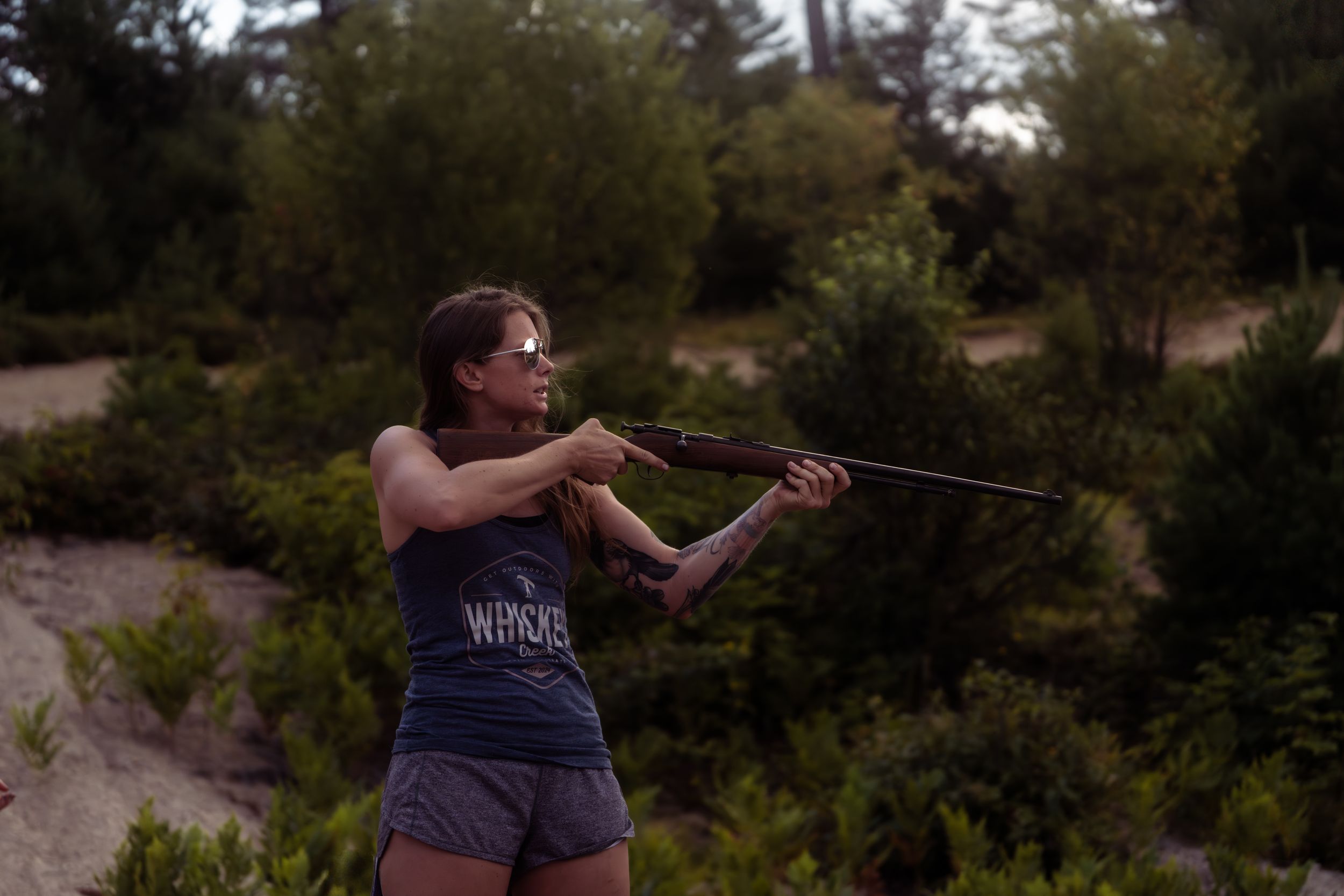 A person wearing a gray shirt aims a rifle while practicing target shooting in a wooded outdoor setting.