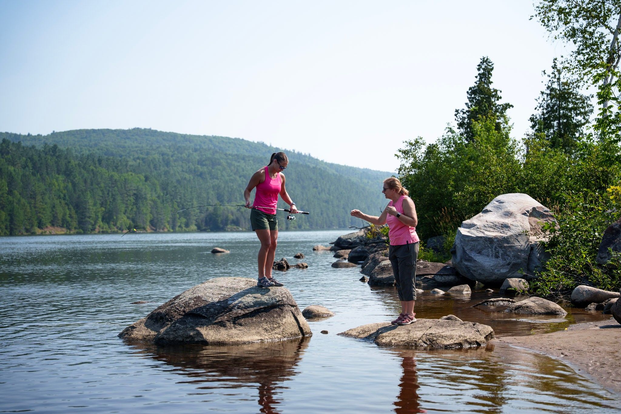 People enjoying a sunny summer day on large rocks by a peaceful lake surrounded by forested hills.
